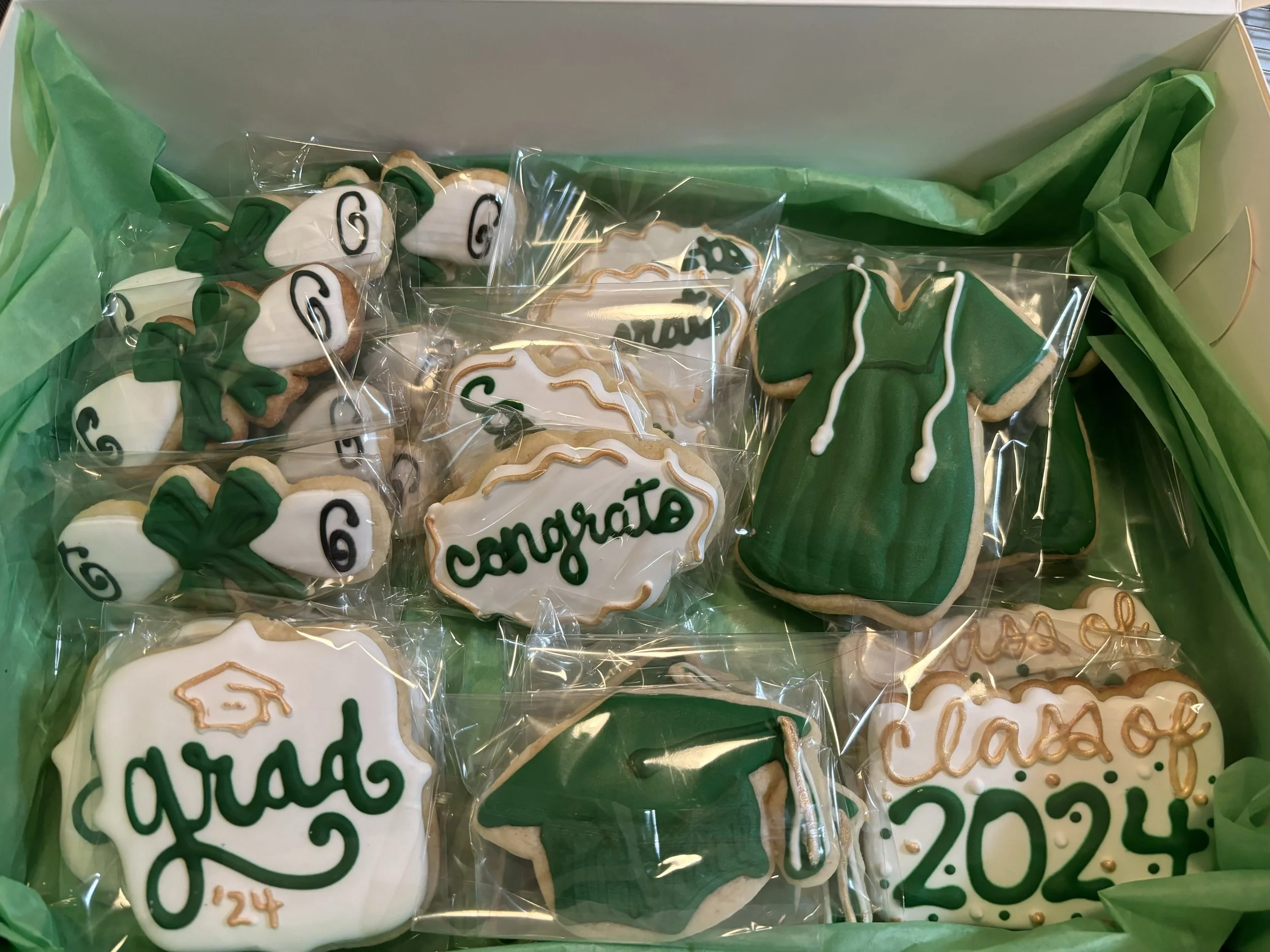 A box of decorated high school graduation cookies, including caps, gowns, diplomas, and congratulatory messages, all wrapped in clear plastic.