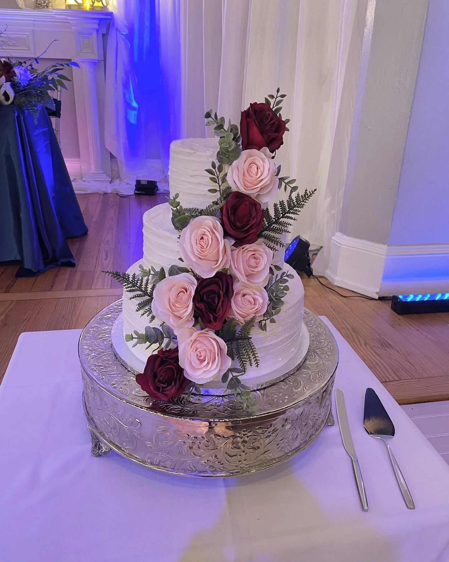 Three-tier wedding cake decorated with pink and deep red roses and greenery, placed on a decorative silver cake stand with a cake knife and server beside it.
