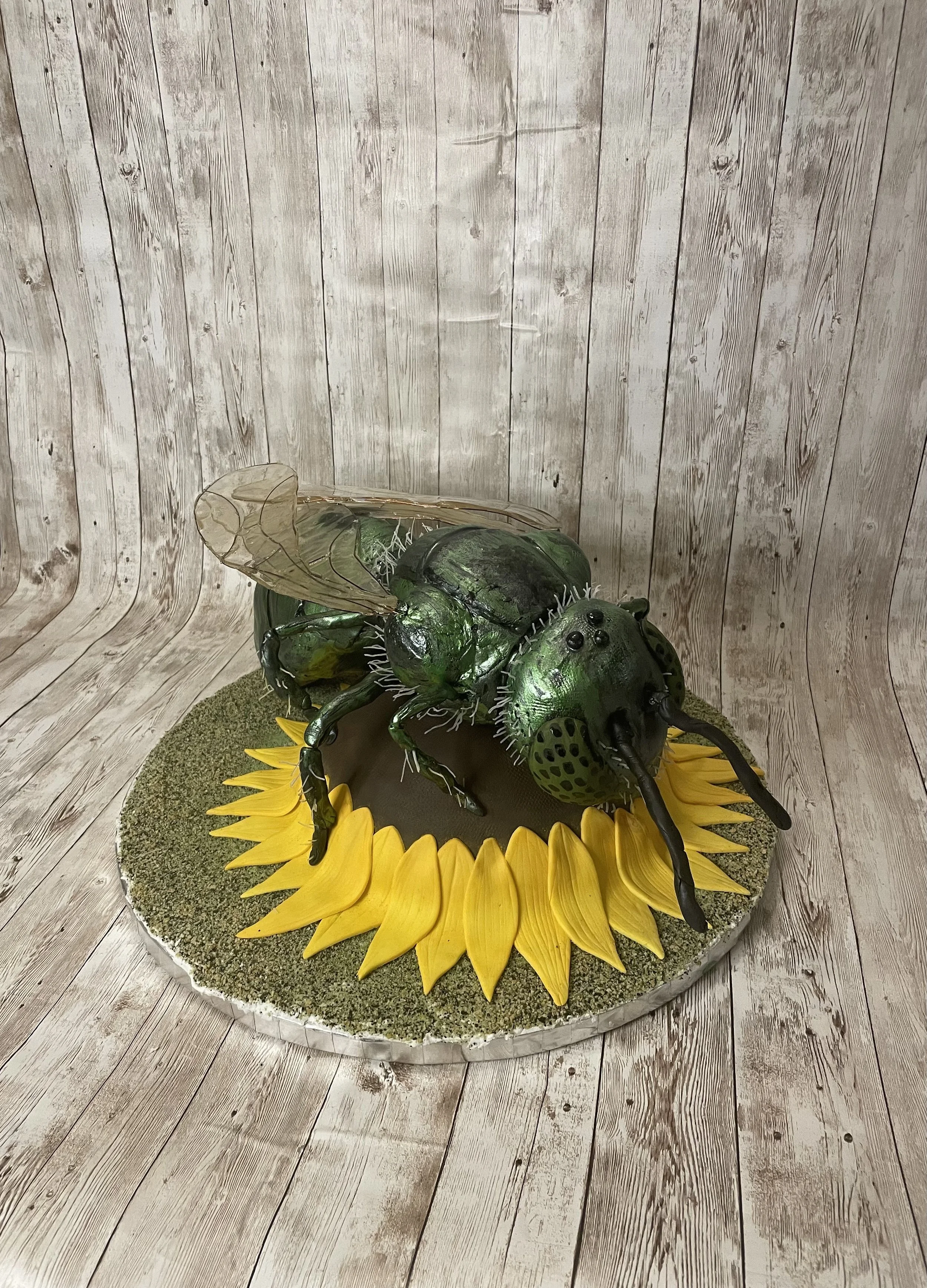 A cake sculpture of a bee with black antennae and large eyes, placed on a sunflower-shaped base with yellow petals, on a round edible grass base, set against wood-paneled background.