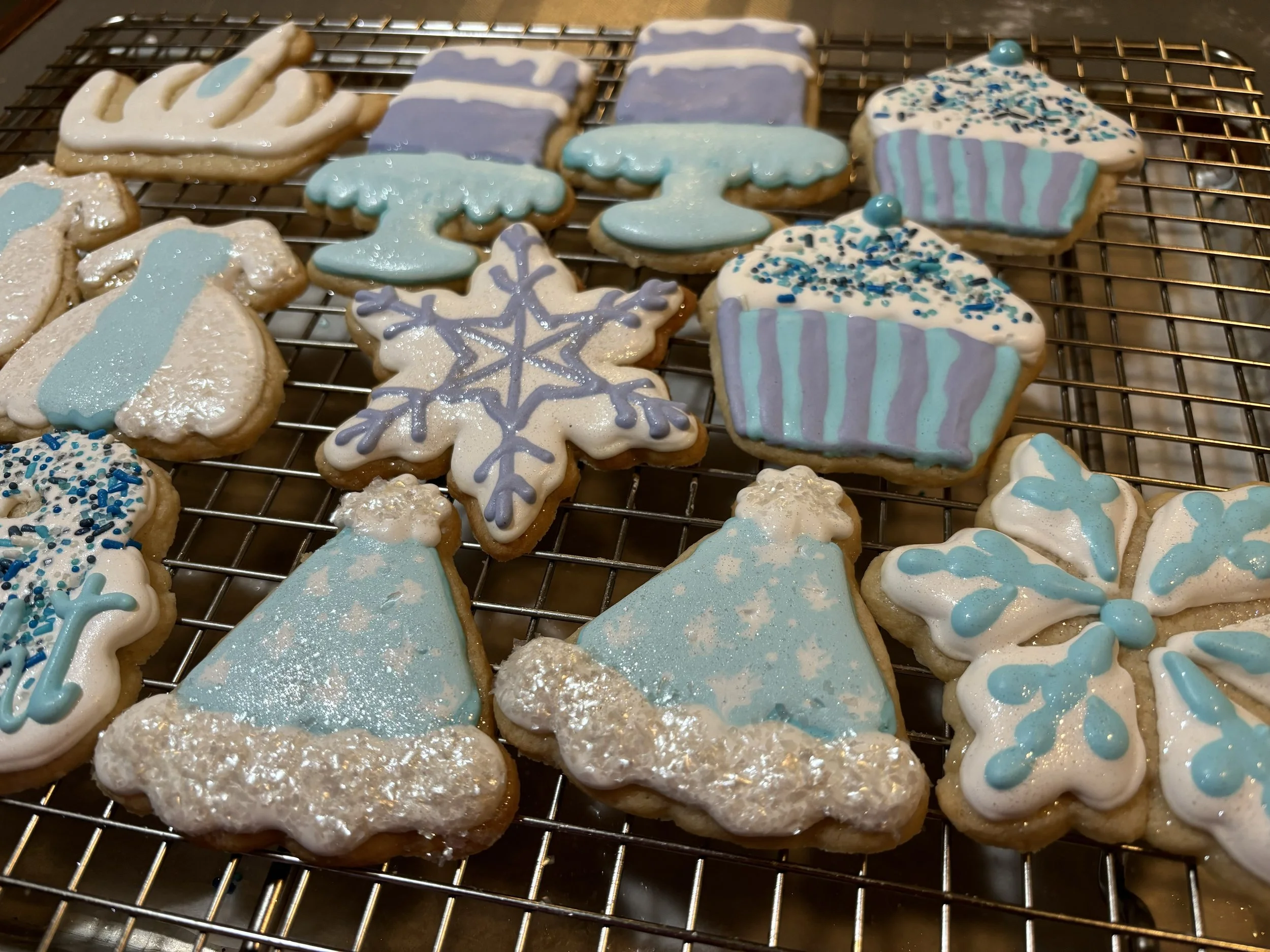 A variety of decorated birthday cookies on a cooling rack, including snowflakes, hats, and cupcakes, decorated in blue, white, and purple icing with sprinkles.
