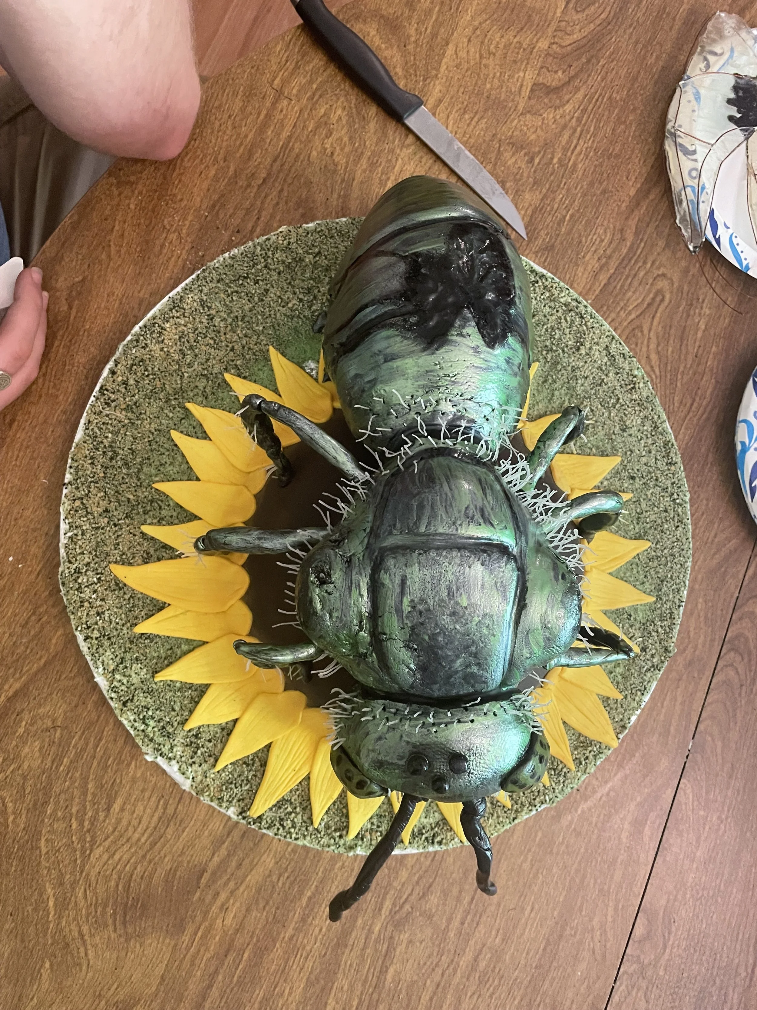 A cake designed to look like a green metallic sweat bee, decorated with green and black colors, surrounded by yellow edible petals, on a round cake board.