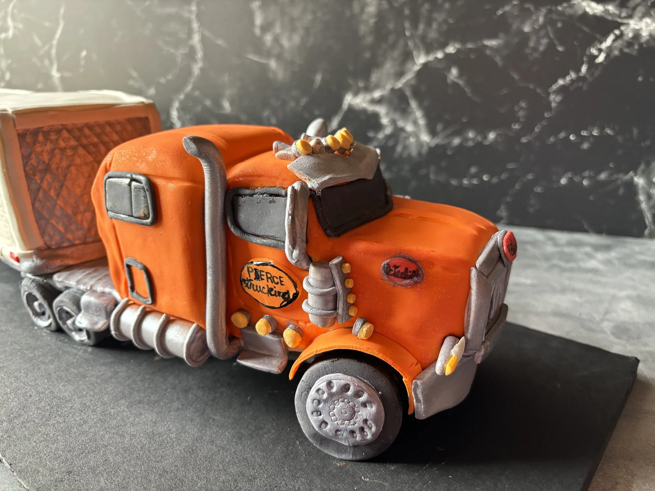 Decorative cake designed to resemble a semi-truck, with orange icing, gray details, and black wheels, displayed on a black base.