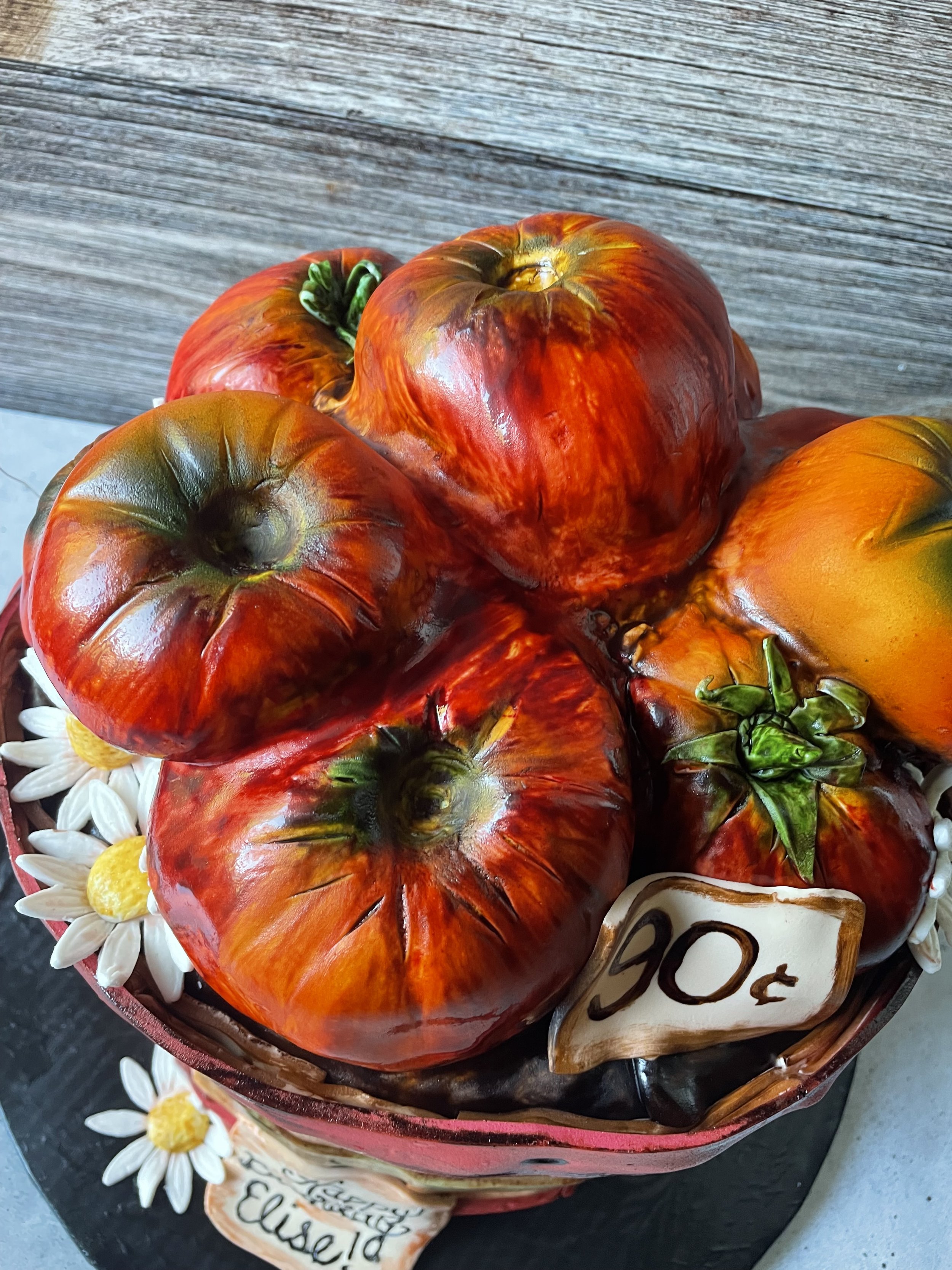 A basket of large, ripened heirloom tomatoes with a handwritten price tag of 90 cents attached, with white and yellow daisies.