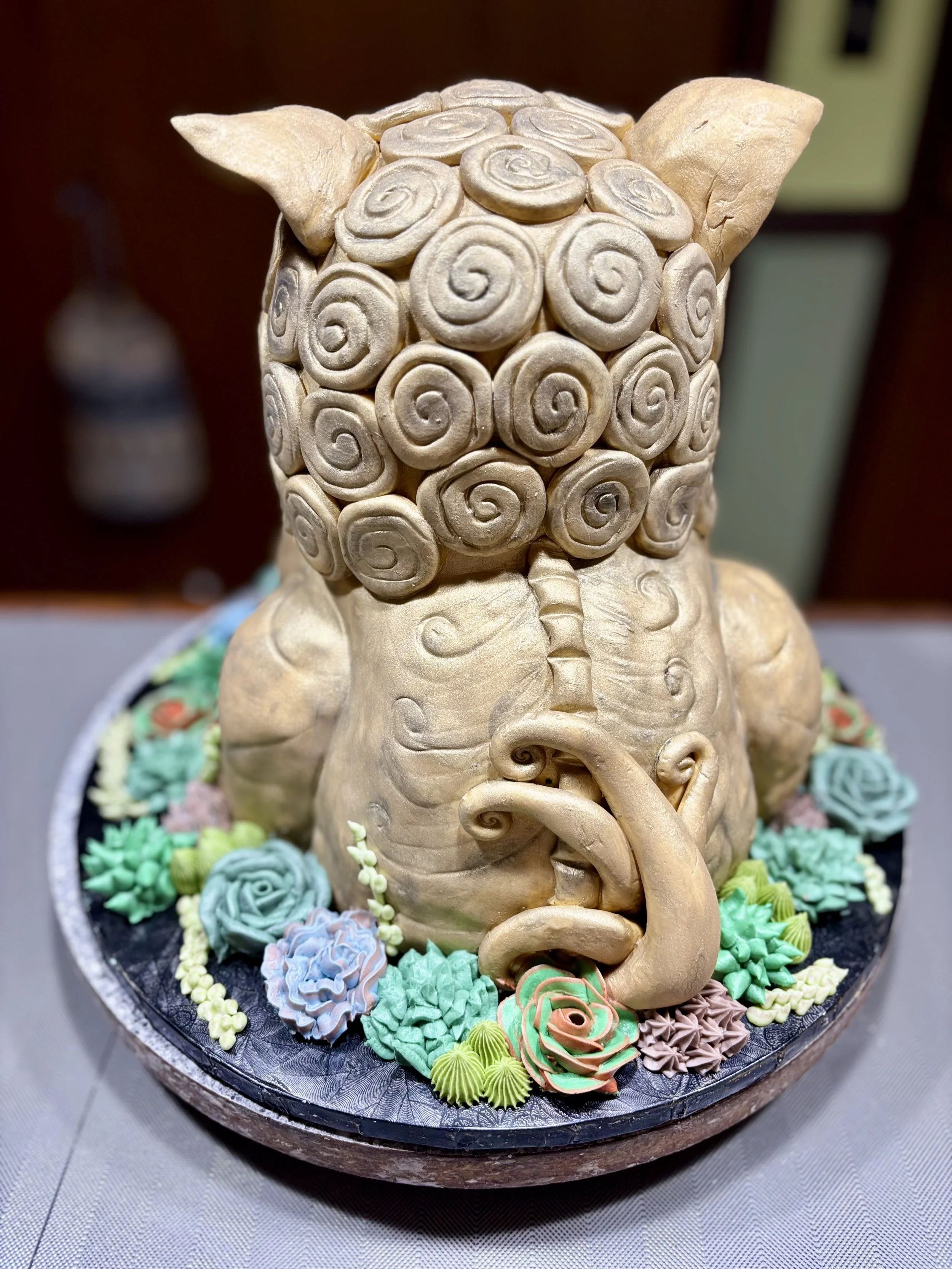 A cake decorated to look like a fu dog with spiral-patterned hair, surrounded by colorful succulent icing decorations.
