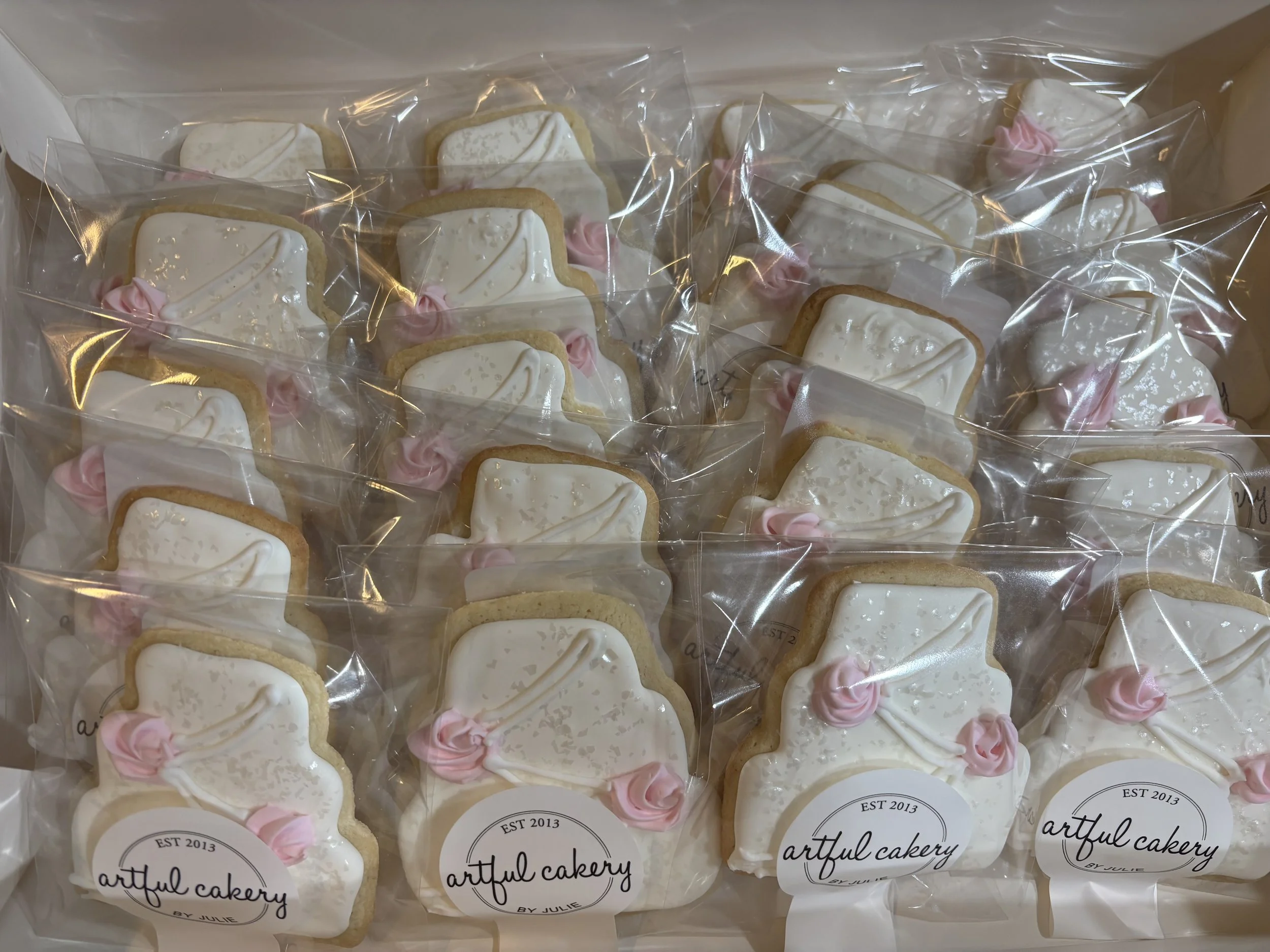 Packaged sugar cookies with white icing and pink accents, labeled 'artful cakery by Julie'.