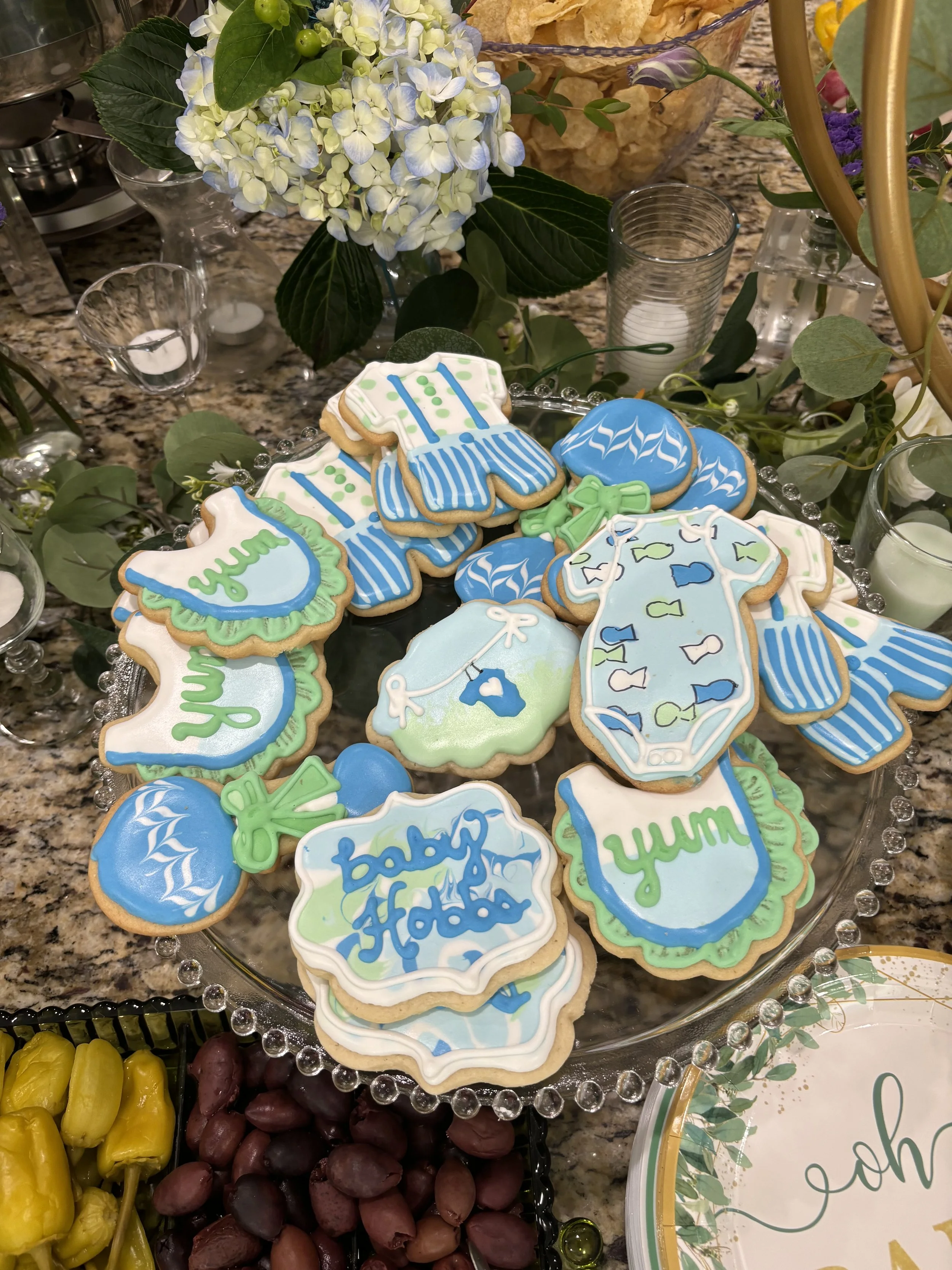 Decorative baby shower cookies with baby clothing, bottles, and bib designs, placed on a clear glass platter with floral decorations and surrounded by candles and snacks.
