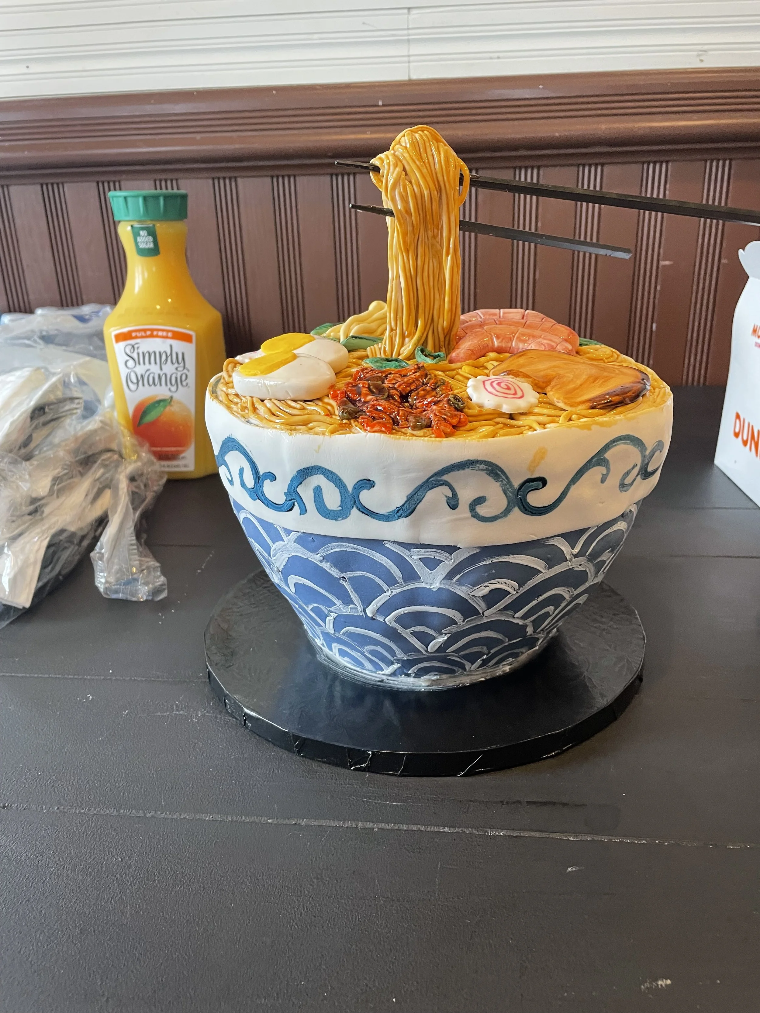 Cake designed to look like a bowl of ramen with noodles, a boiled egg, slices of meat, vegetables, and a narutomaki, decorated with blue wave patterns, placed on a black circular base.