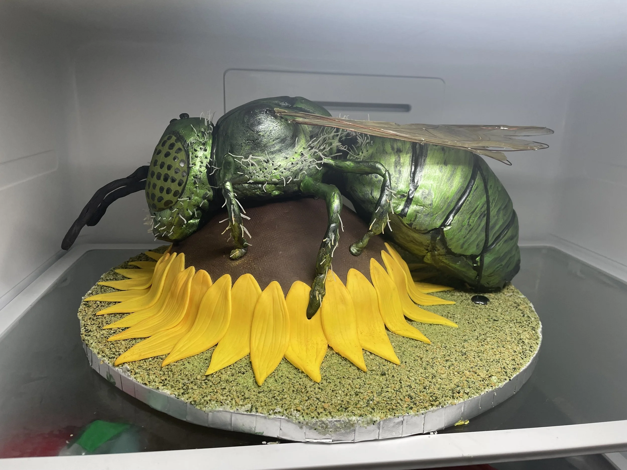 green metallic sweat bee cake on an edible sunflower decorated cake inside a refrigerator