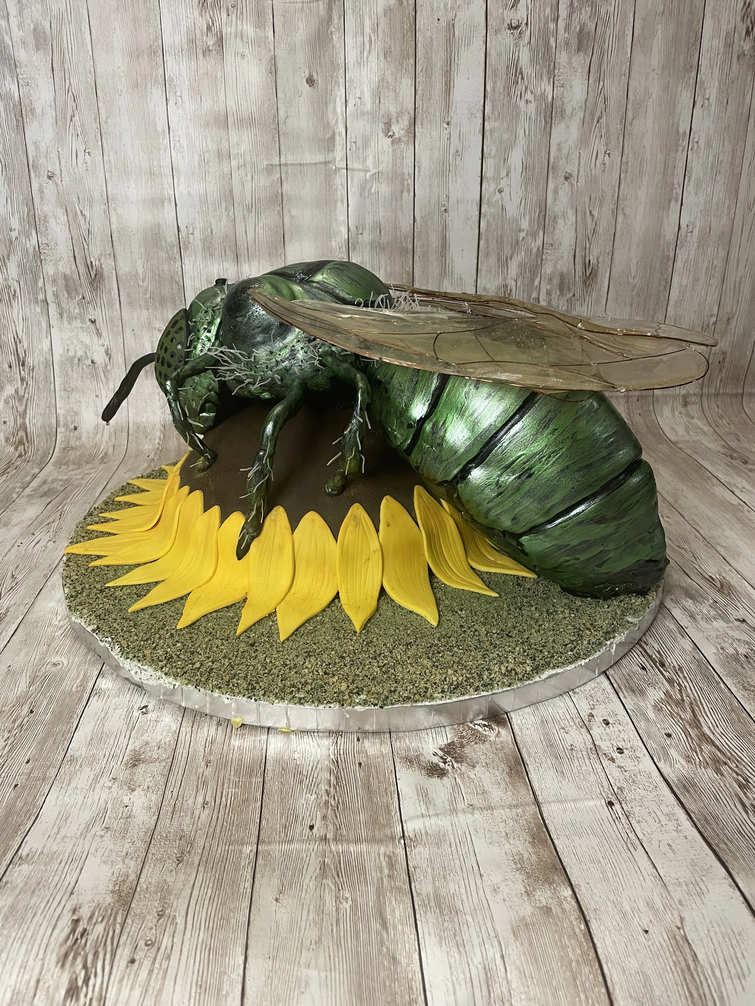 A green metallic sweat bee cake decorated with transparent wings resting on a yellow sunflower, set on a round base with moss-like decoration, against a wooden plank background.