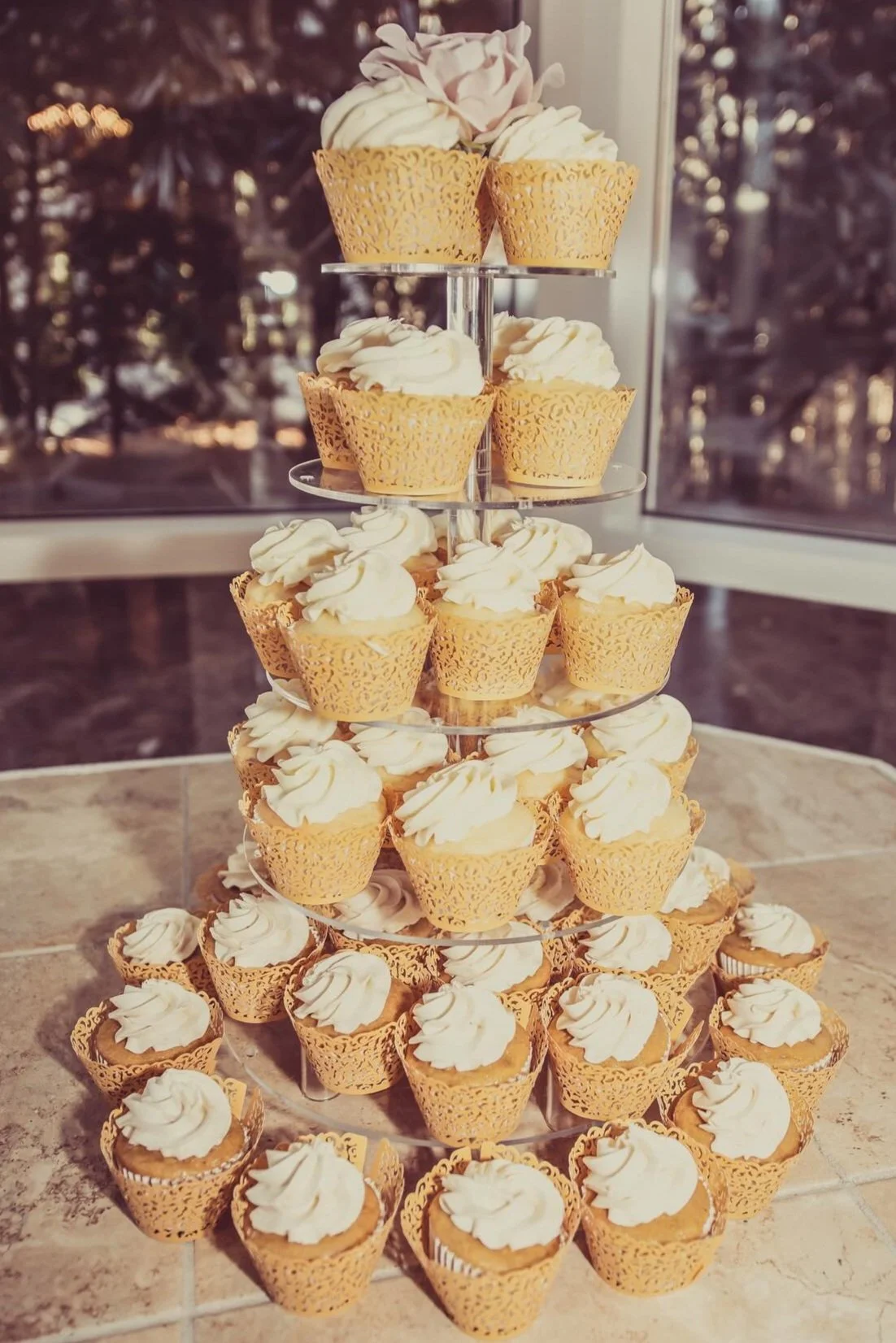 A tiered display of cupcakes with white frosting in gold lace-like wrappers on a tiled table near a window, with trees visible outside.