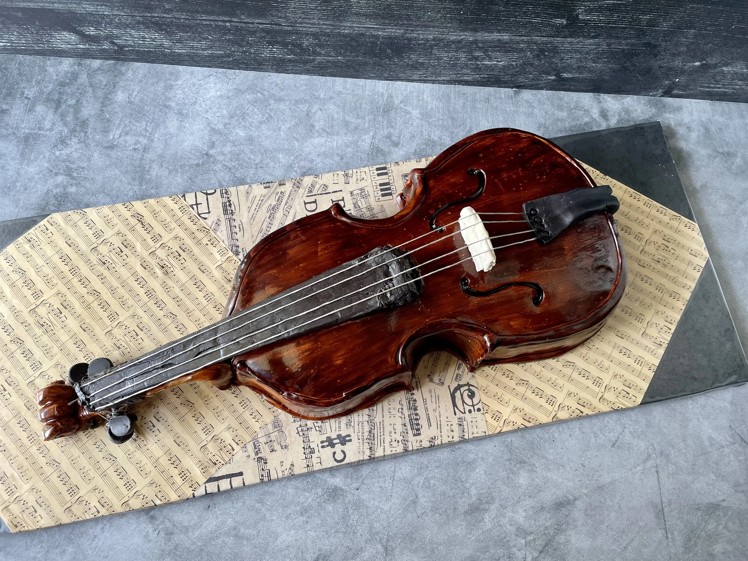 A cake shaped like a violin, decorated with wood grain finish, placed on sheet music paper.