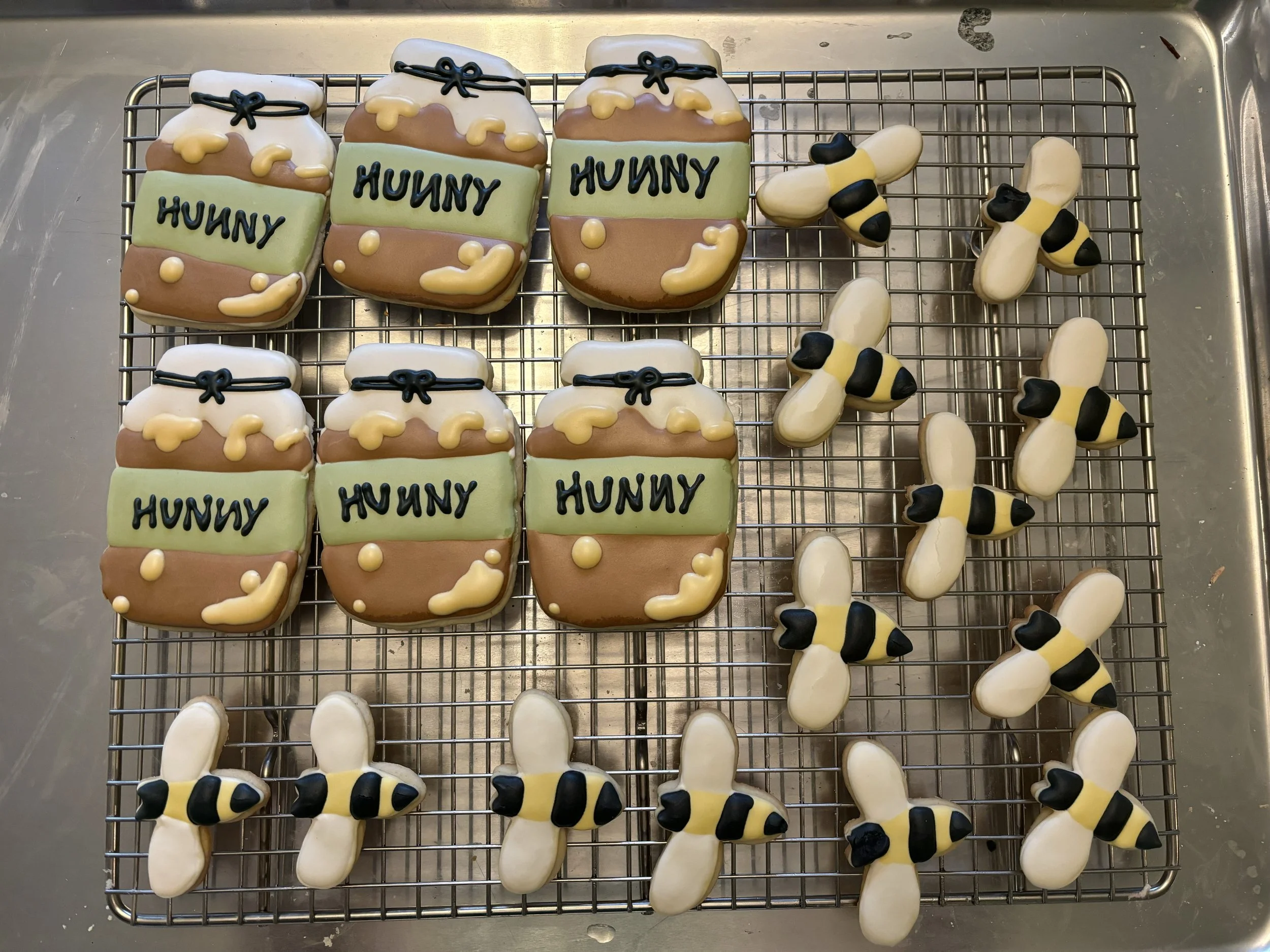 Decorated sugar cookies shaped like honey jars and bees on a wire rack. The honey jar cookies have the word 'HUNNY' written on them and are decorated with yellow, brown, white, and black icing. The bee cookies are decorated with black and yellow icing to resemble bees.