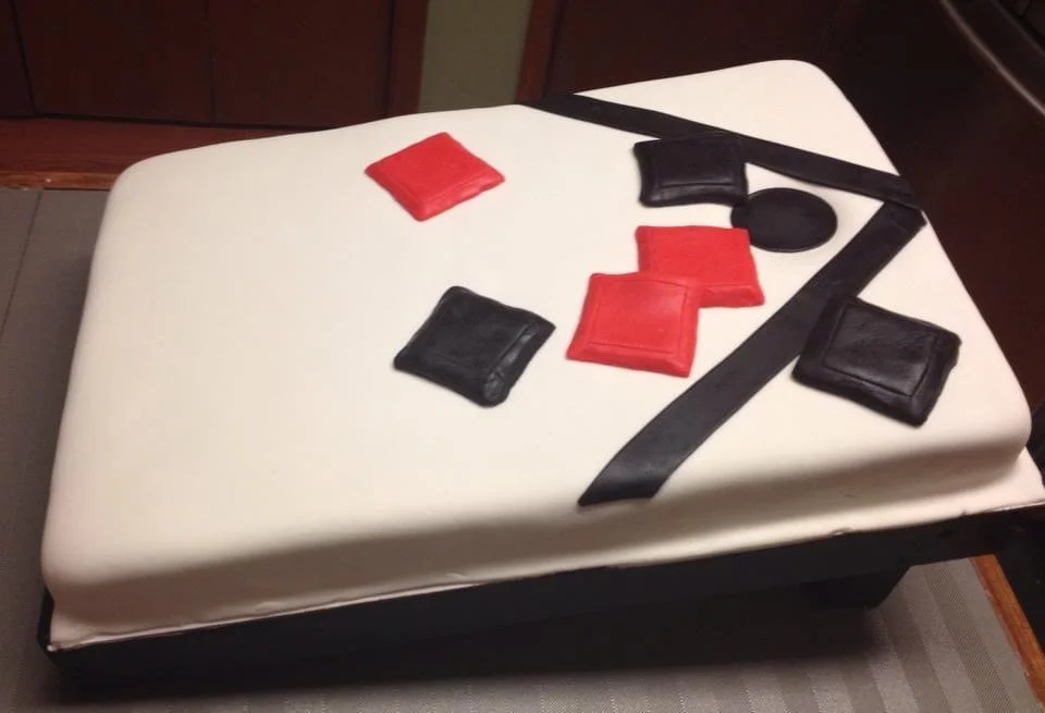 Rectangular cake with white fondant decorated with black and red squares, a black circle, and black strips resembling a corn hole game