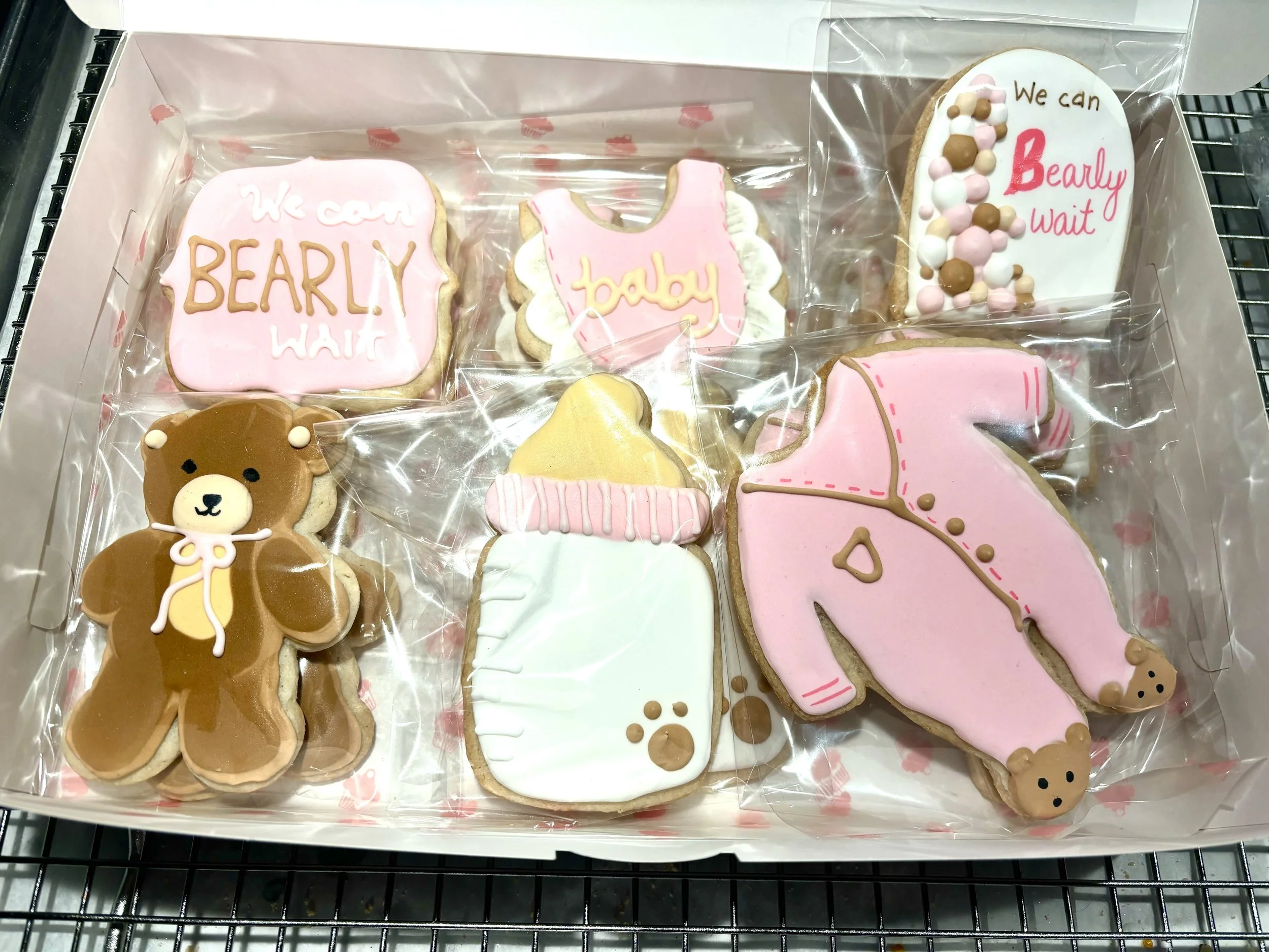 A box of decorated cookies with baby and bear themes, including a bear-shaped cookie, a pink baby onesie, a baby bottle, a pink elephant, and decorated cookies with messages like 'We can Bearly Wait,' all wrapped in clear plastic.