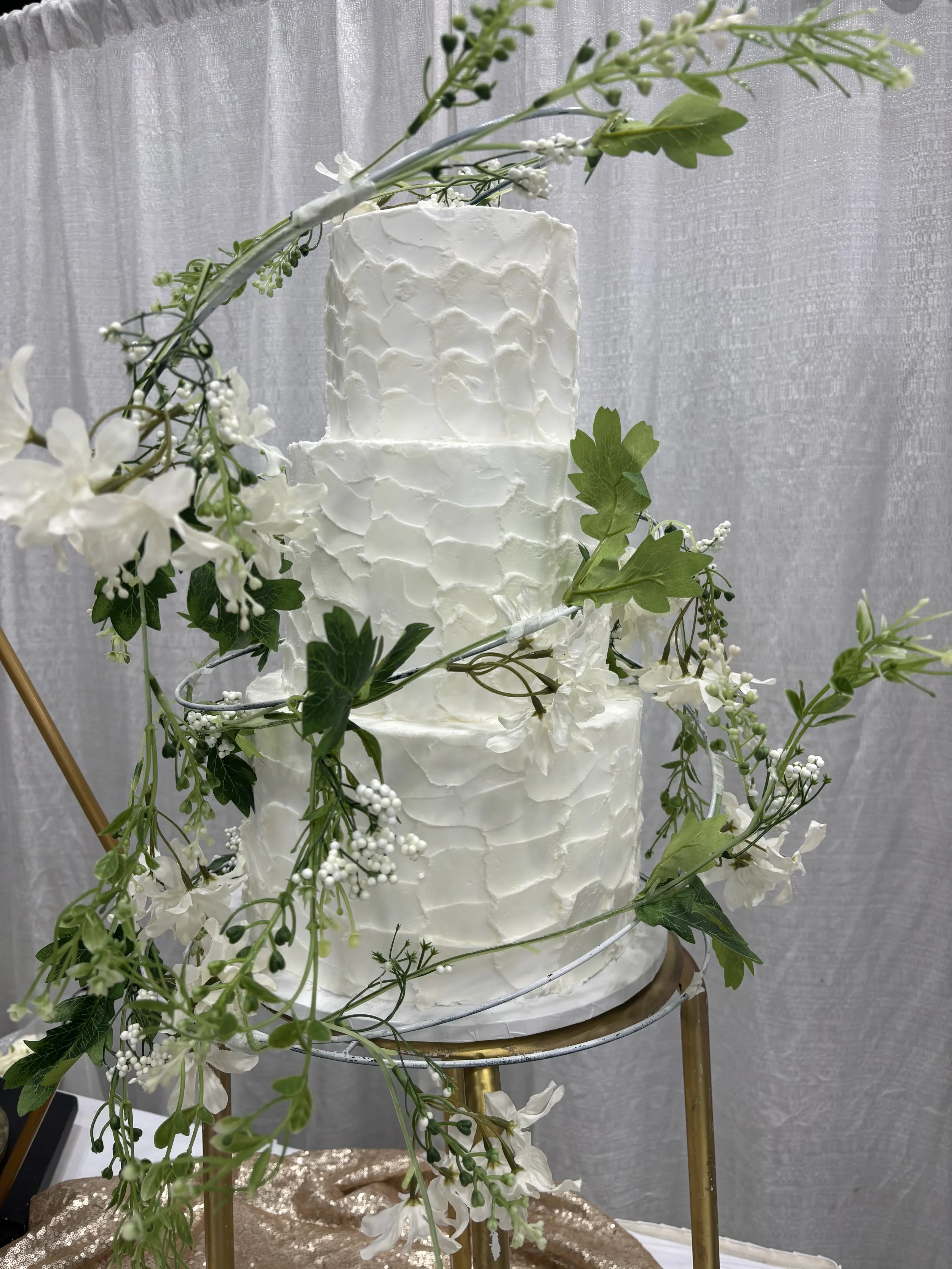 Three-tier white wedding cake decorated with white flowers and green leaves, surrounded by floral garland.