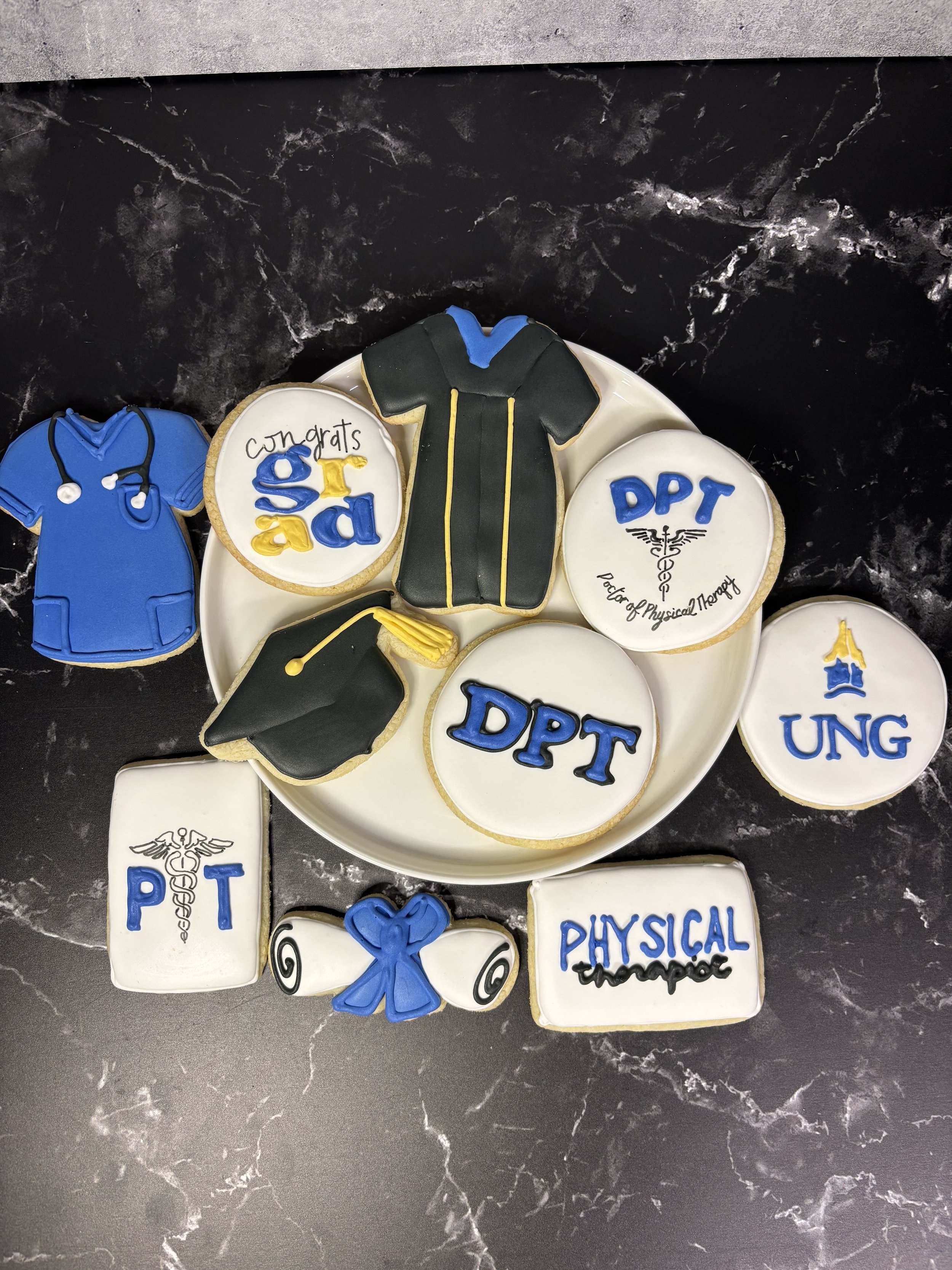 Cookies decorated with themes related to physical therapy education, including graduation caps, medical symbols, and related slogans on a black marble surface.
