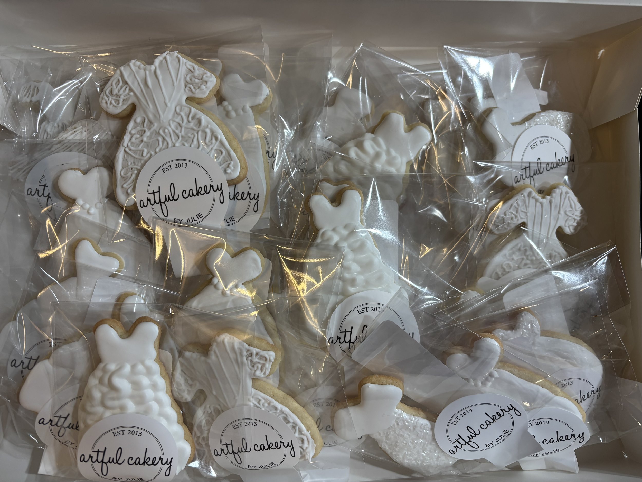 Assorted white sugar cookies decorated with icing, shaped like wedding dresses  individually wrapped in clear plastic, with labels from Artful Cakery.