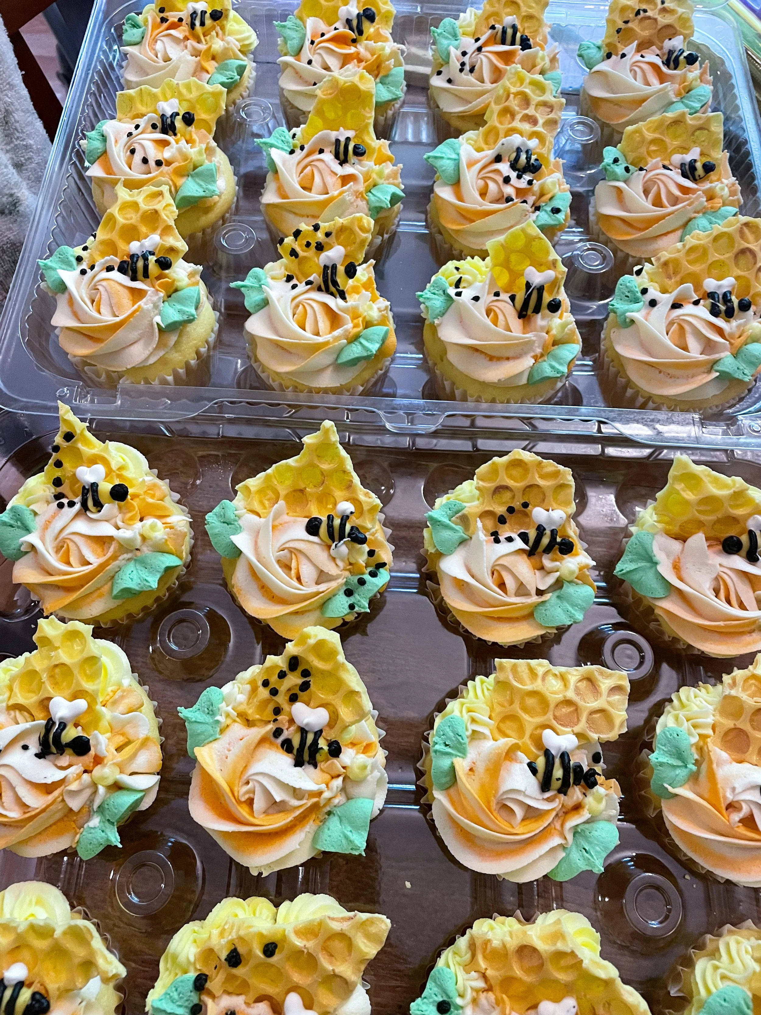 Decorative honey bee-themed cupcakes with swirl frosting, green leaf decorations, and honeycomb-shaped toppers arranged in rows on clear plastic trays.