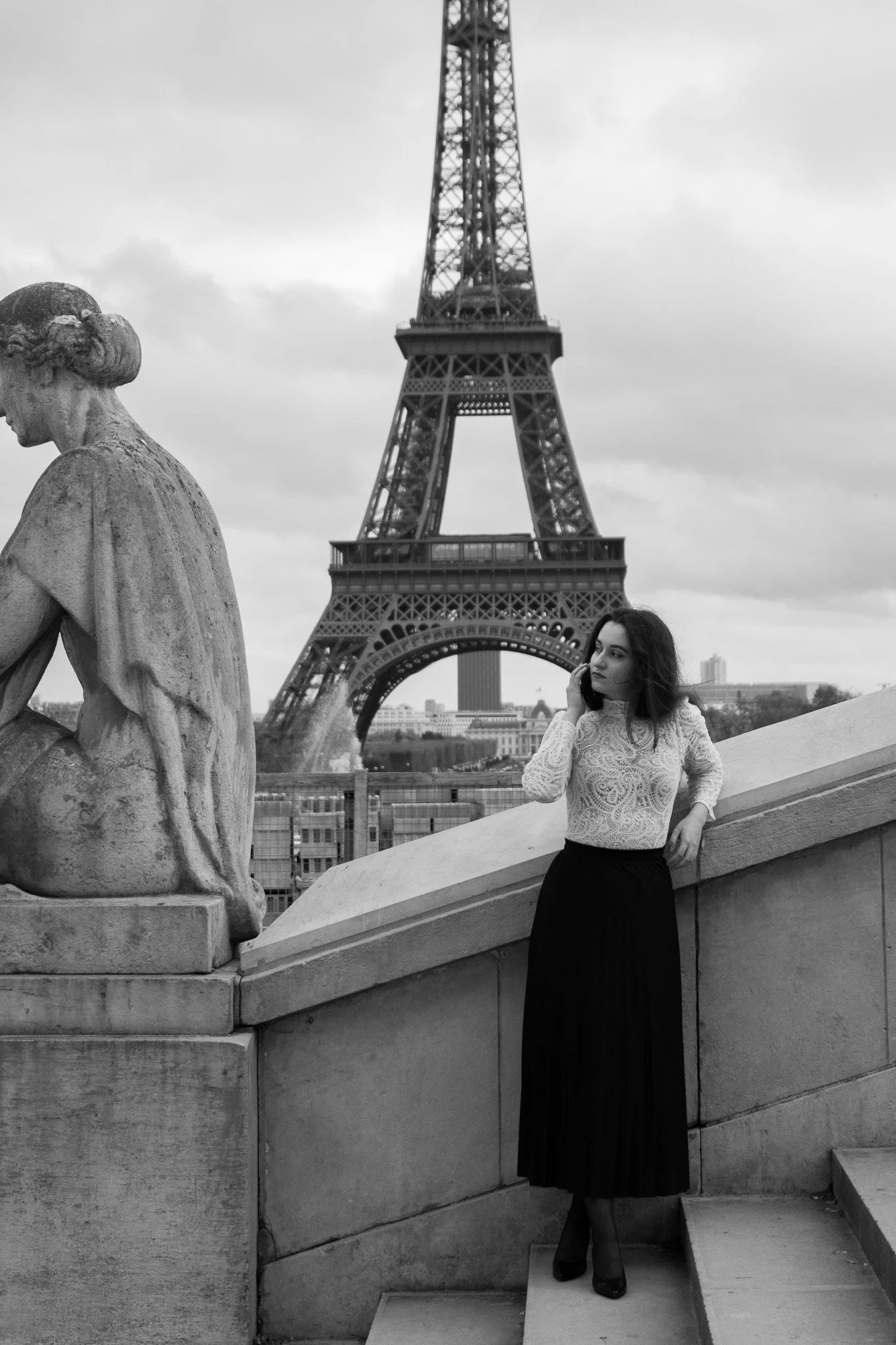A woman in a lace top and long skirt stands on stairs in front of a statue, talking on her phone, with the Eiffel Tower in the background.