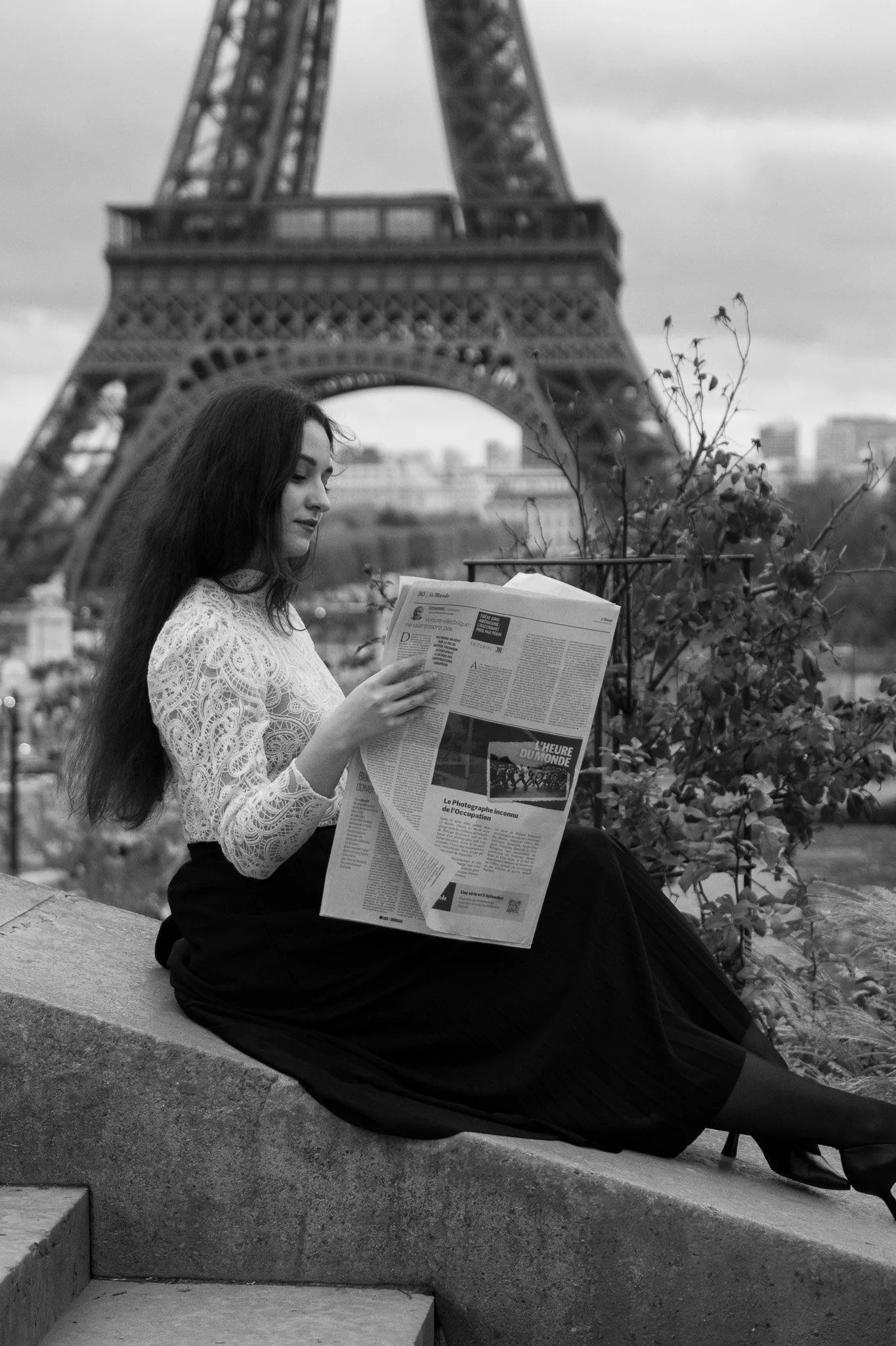A woman with long hair wearing a lace top and a skirt, sitting on a stone ledge and reading a newspaper with the Eiffel Tower in the background.