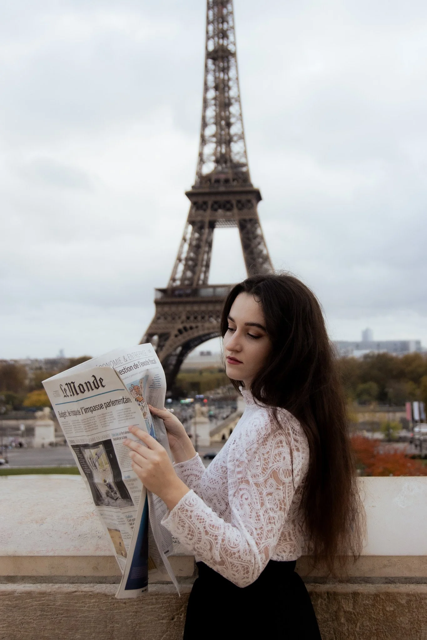 A young woman with long brown hair and a white lace top reads a newspaper while standing in front of the Eiffel Tower in Paris, France.