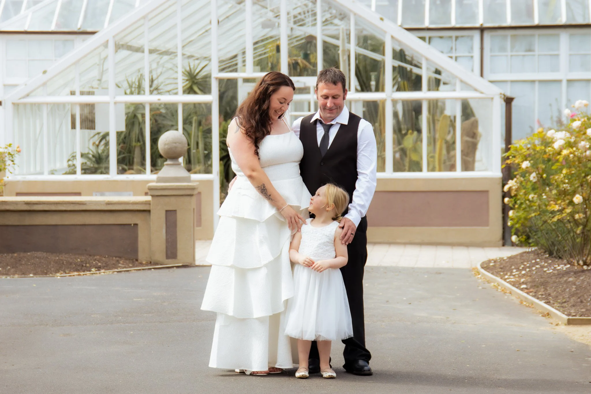 A family standing outdoors near a greenhouse. An adult woman, adult man, and a young girl are smiling and looking at each other. The woman is wearing a white dress, the man is in a black vest and white shirt, and the girl is in a white dress.