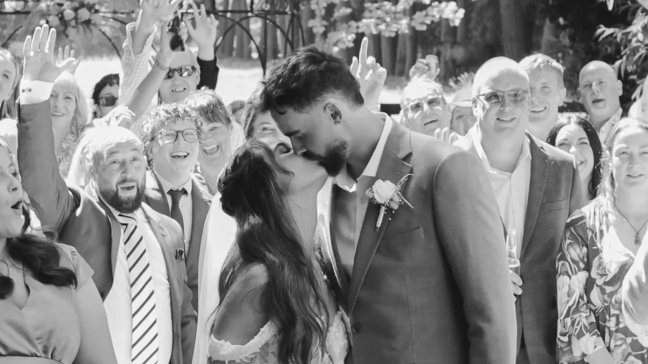 Black and white photo of a wedding kiss between a woman in a lace dress and a man in a suit with a boutonnière, surrounded by smiling guests.