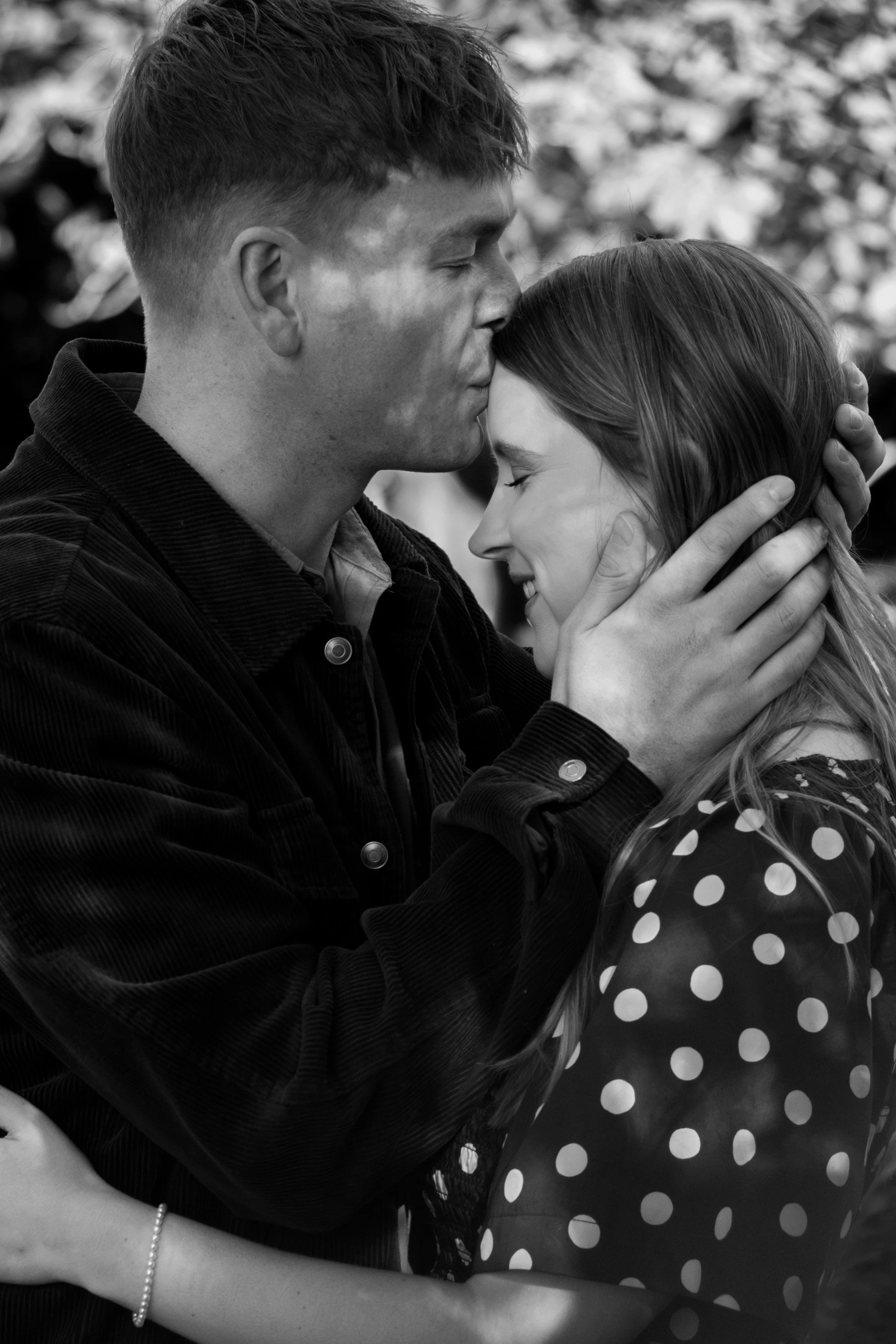 Black and white photo of a wedding kiss between a woman in a lace dress and a man in a suit with a boutonnière, surrounded by smiling guests.