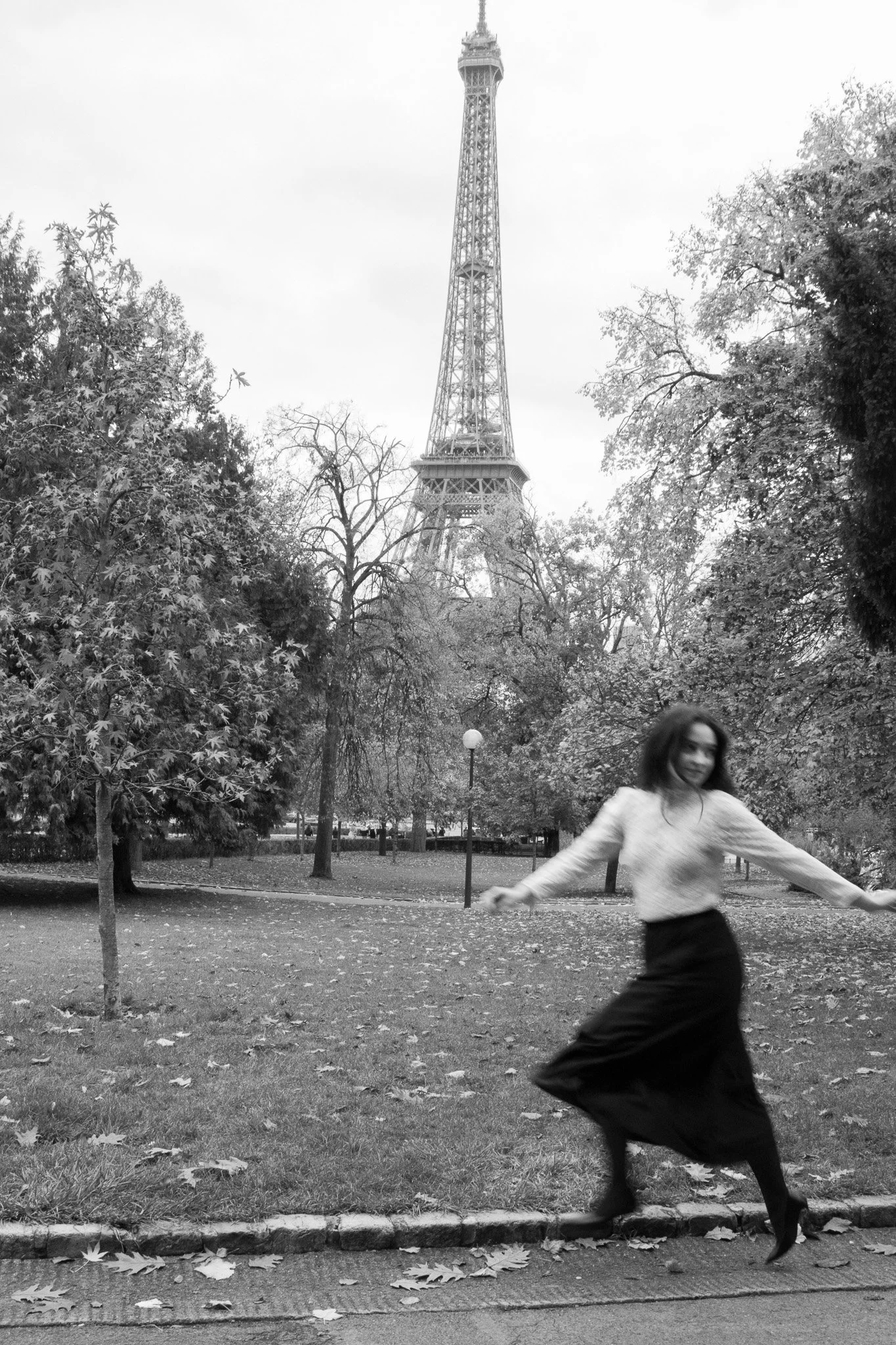 Black and white photo of a woman dancing in a park with the Eiffel Tower in the background.