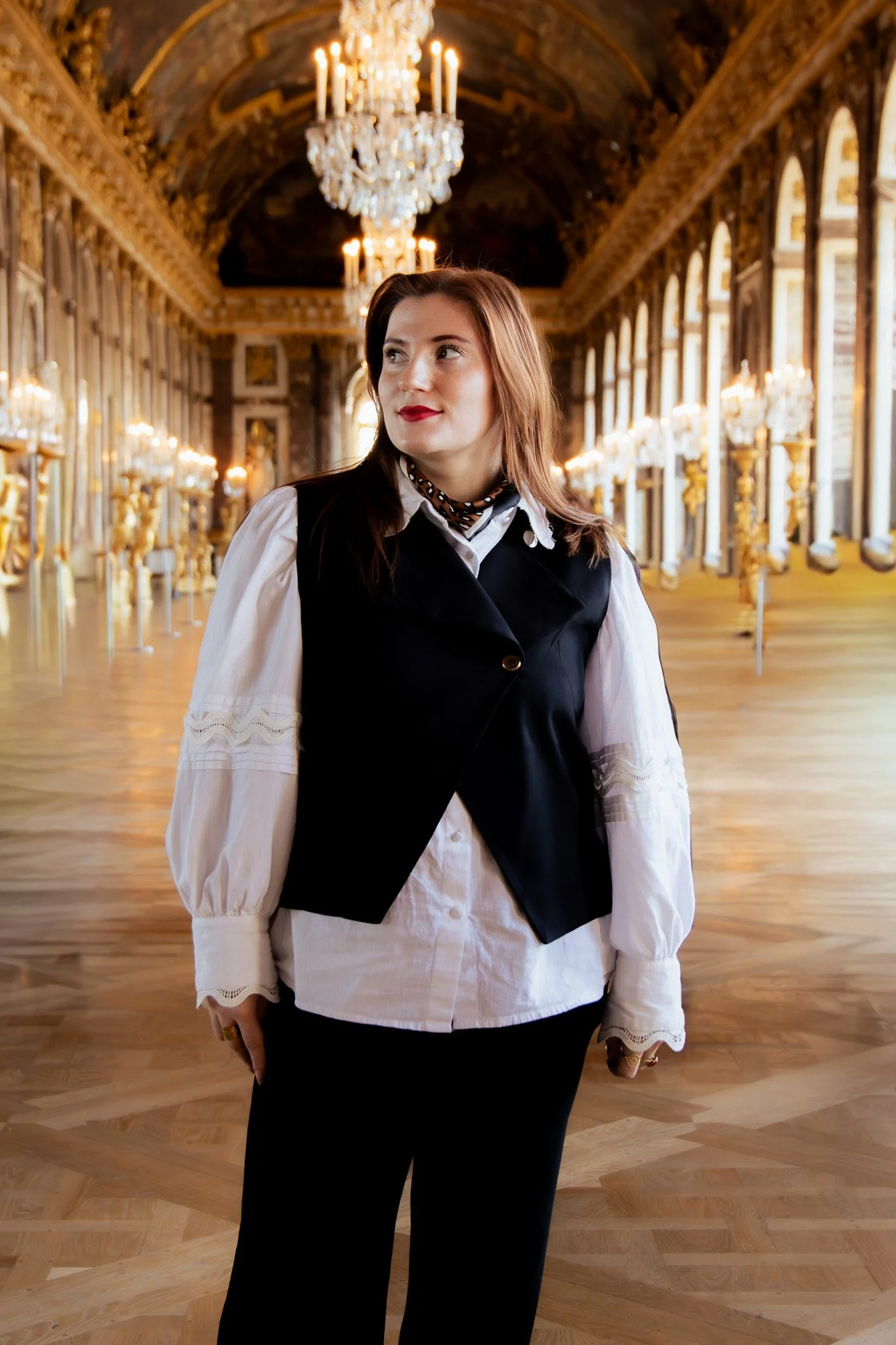 A woman with reddish-brown hair wearing a white blouse, black vest, and black pants standing in an opulent ballroom with ornate chandeliers and gold accents.