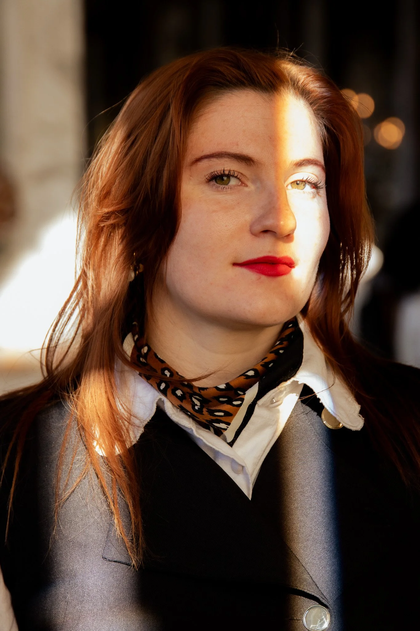 A woman with red hair and green eyes wearing a white shirt, a leopard print scarf, a dark blazer, and red lipstick, in natural sunlight.