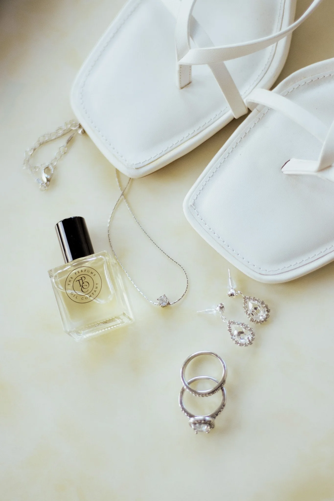 White high-heeled sandals, a small white purse with a chain strap, a bottle of perfume, a necklace with a pendant, a pair of earrings, and three rings on a light-colored surface.