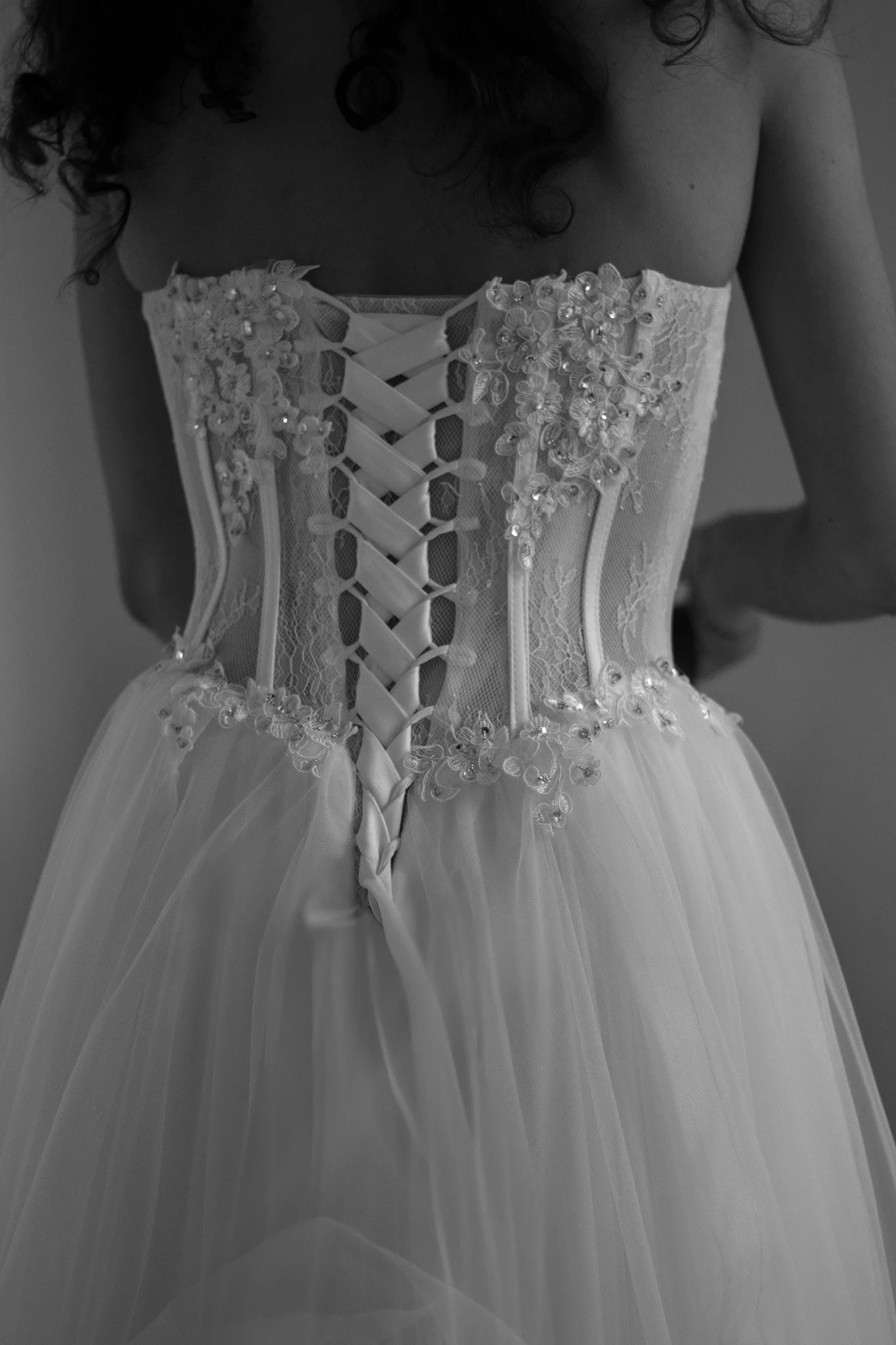 Back view of a woman in an ornate wedding dress with lace and floral embroidery, featuring a corset with ribbon lacing at the back.