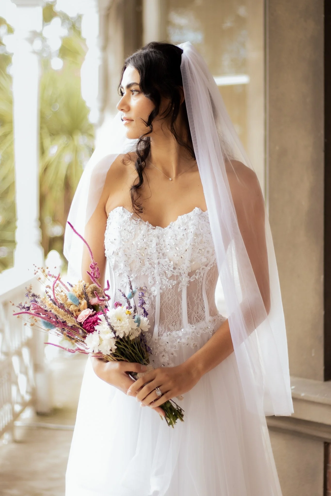 A bride standing indoors in a white wedding dress, holding a colorful bouquet of flowers, with dark hair styled in loose curls and a white veil.