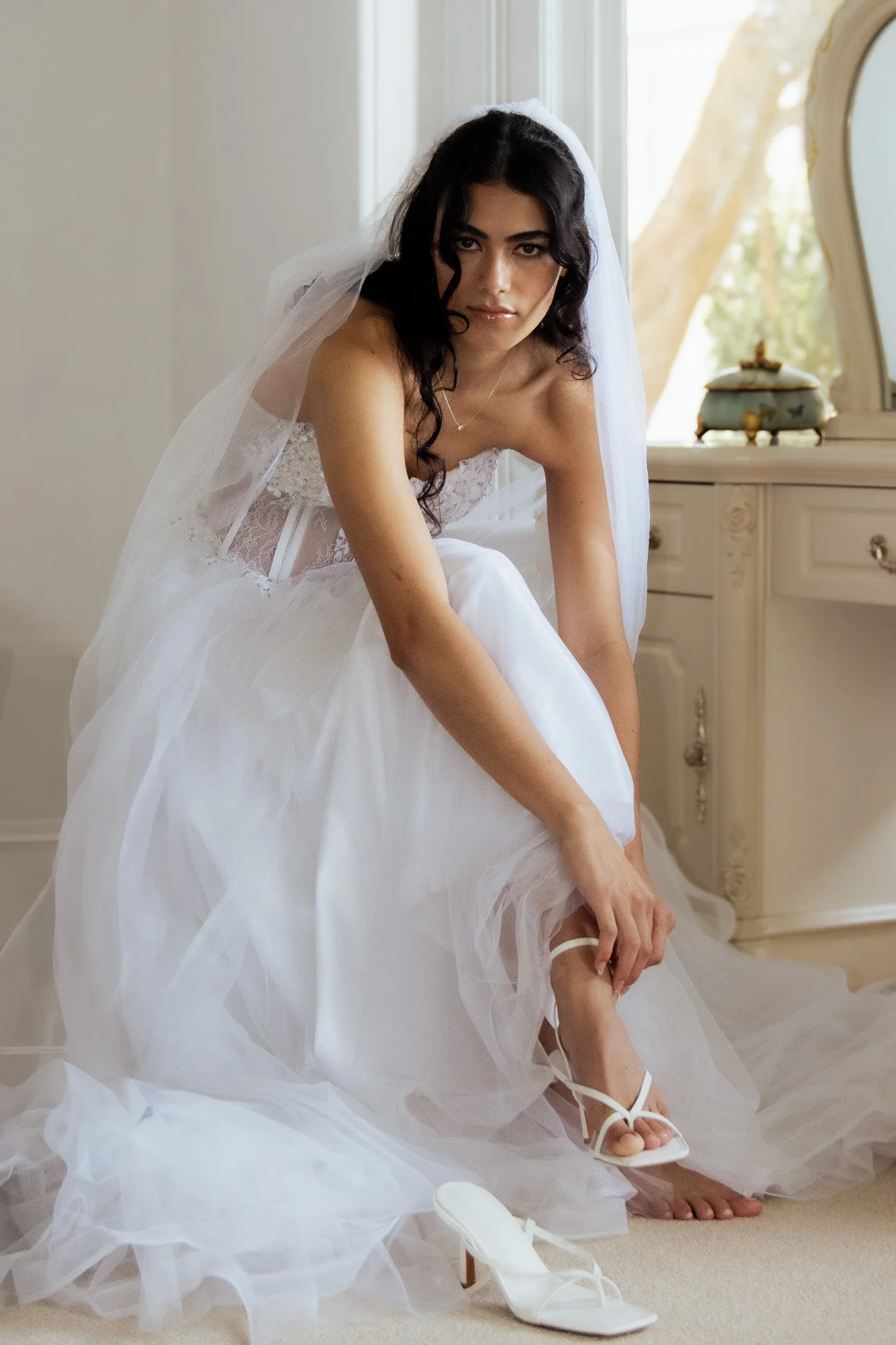 A bride in a white wedding gown is sitting on the floor, adjusting her white high-heeled shoe.