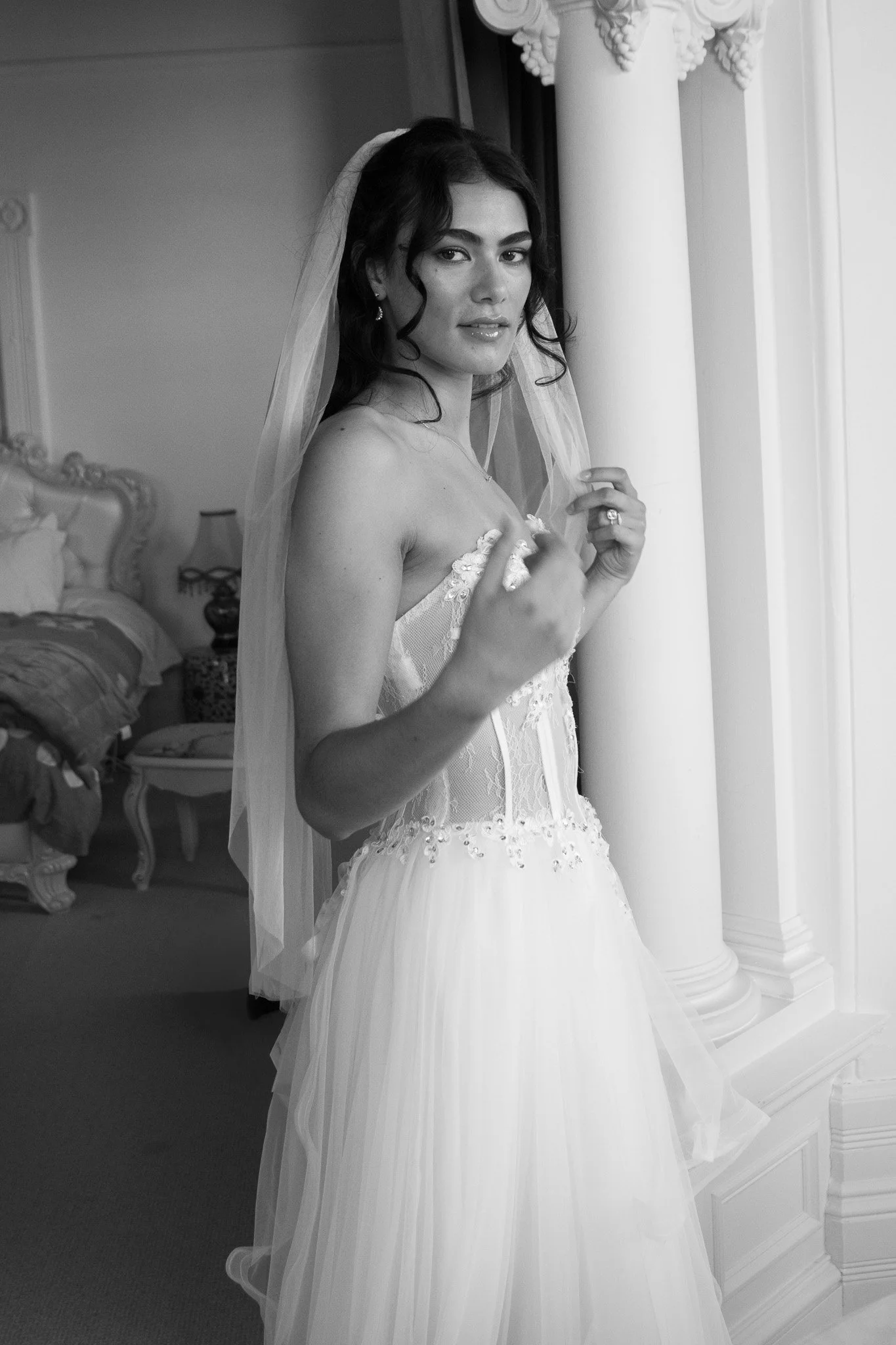 A woman in a wedding dress with a veil standing indoors, near a column, looking at the camera.