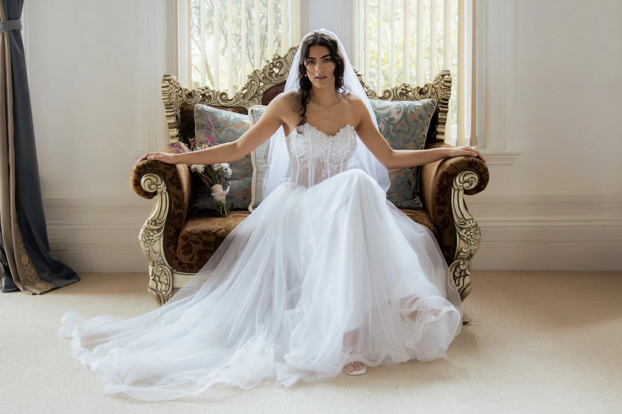 A woman in a white wedding dress sitting on an ornate vintage sofa in a room with light-colored walls and windows.