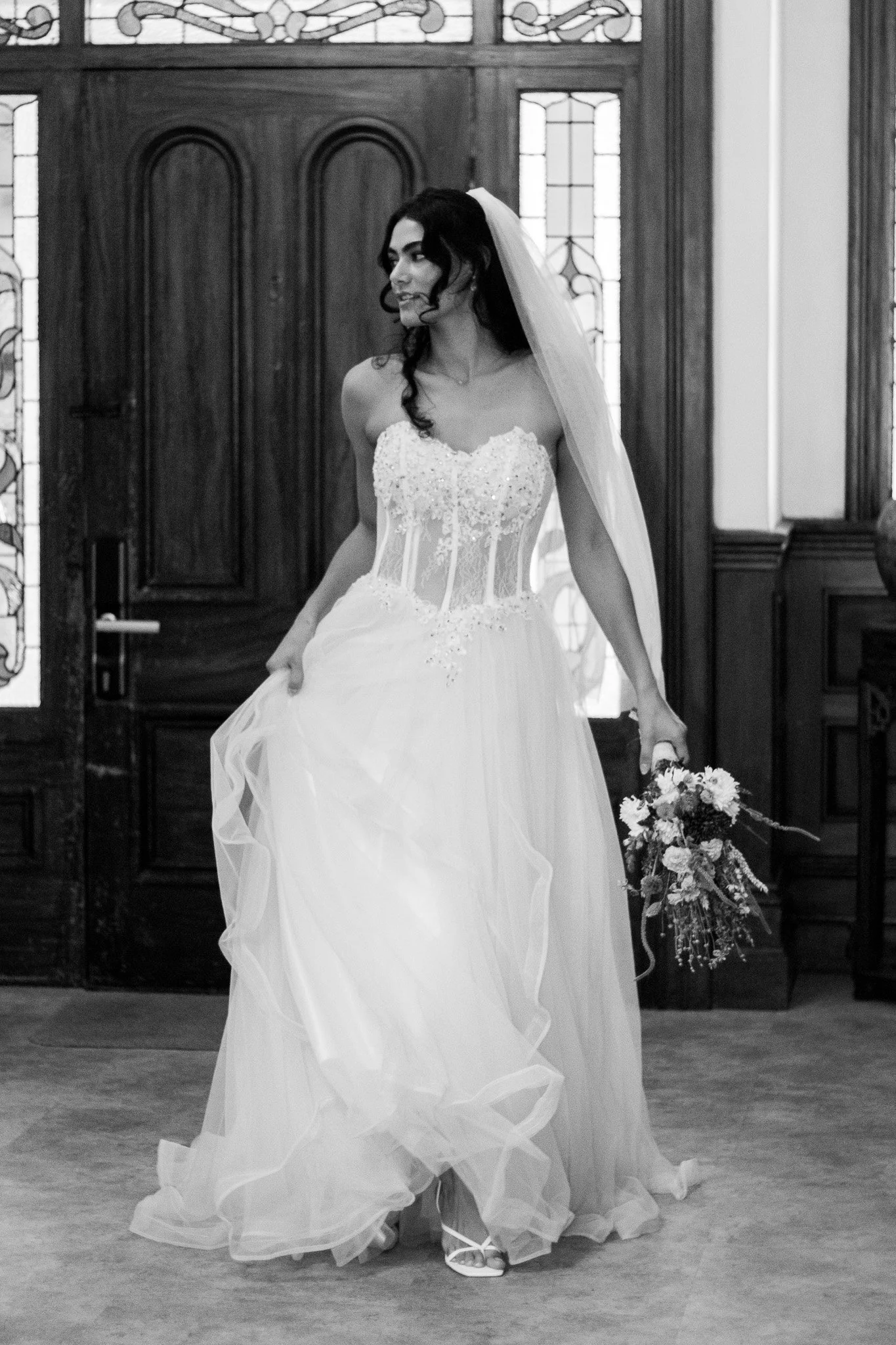 A bride in a wedding dress holding a bouquet, standing indoors near wooden doors and stained glass windows.