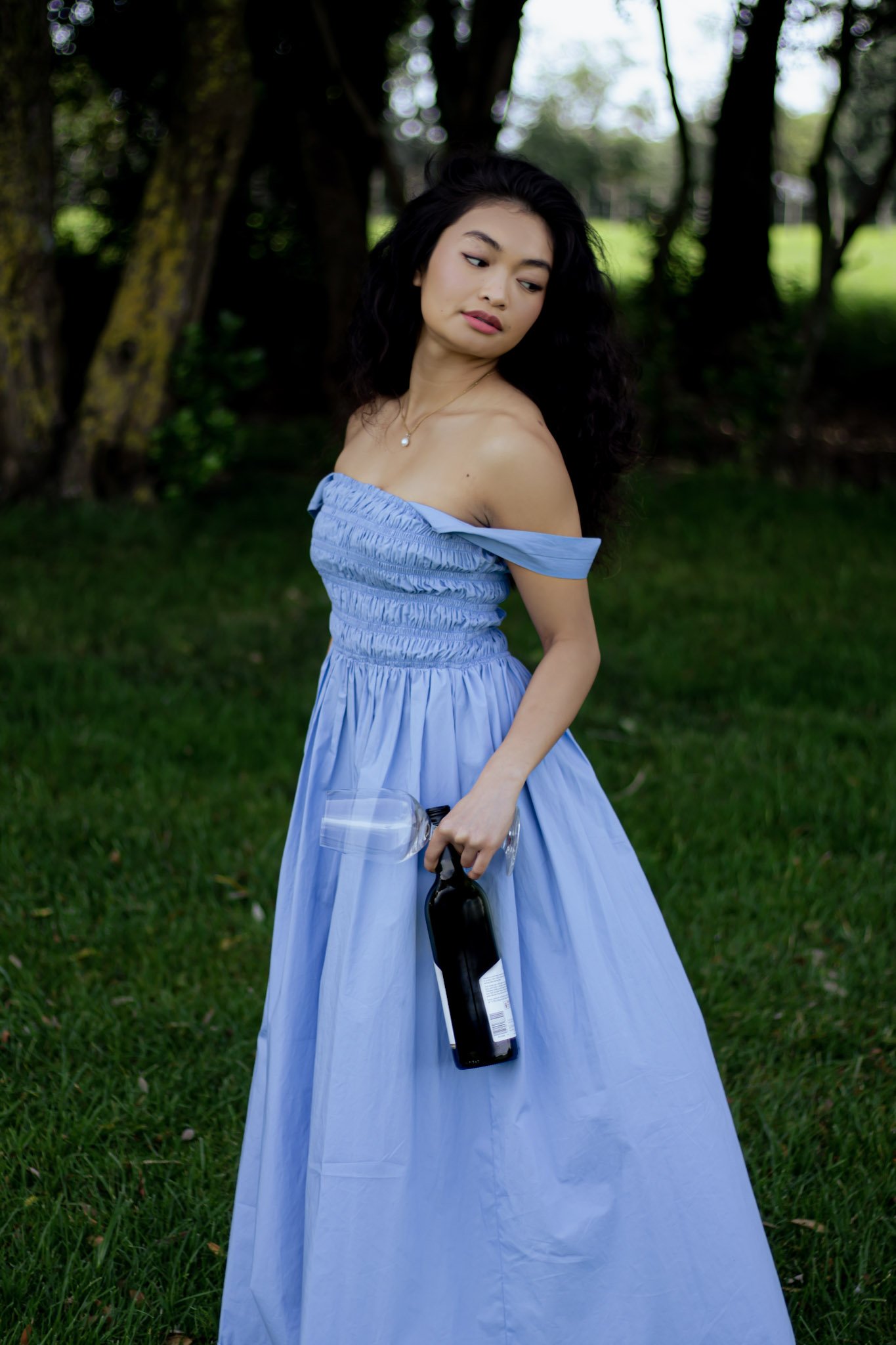 A woman in a light blue off-shoulder dress standing outdoors in a grassy park with trees in the background, holding a plastic water bottle and a dark-colored wine or liquor bottle.
