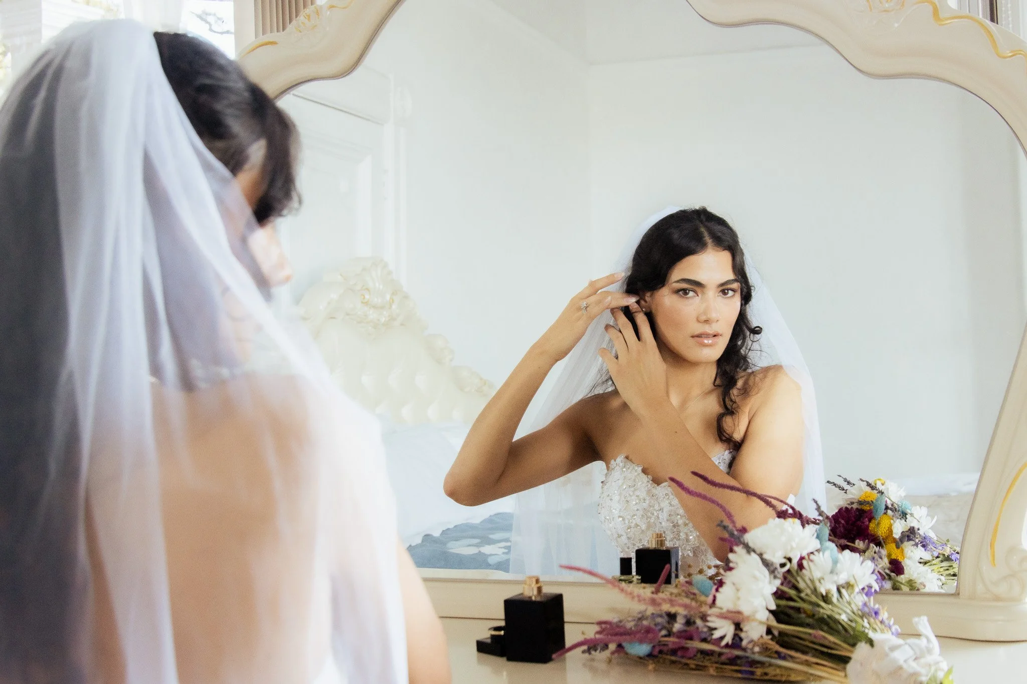 Brunette bride in a wedding dress looking in a mirror as she adjusts her earring, with a bouquet of flowers and perfume bottles on the dresser.