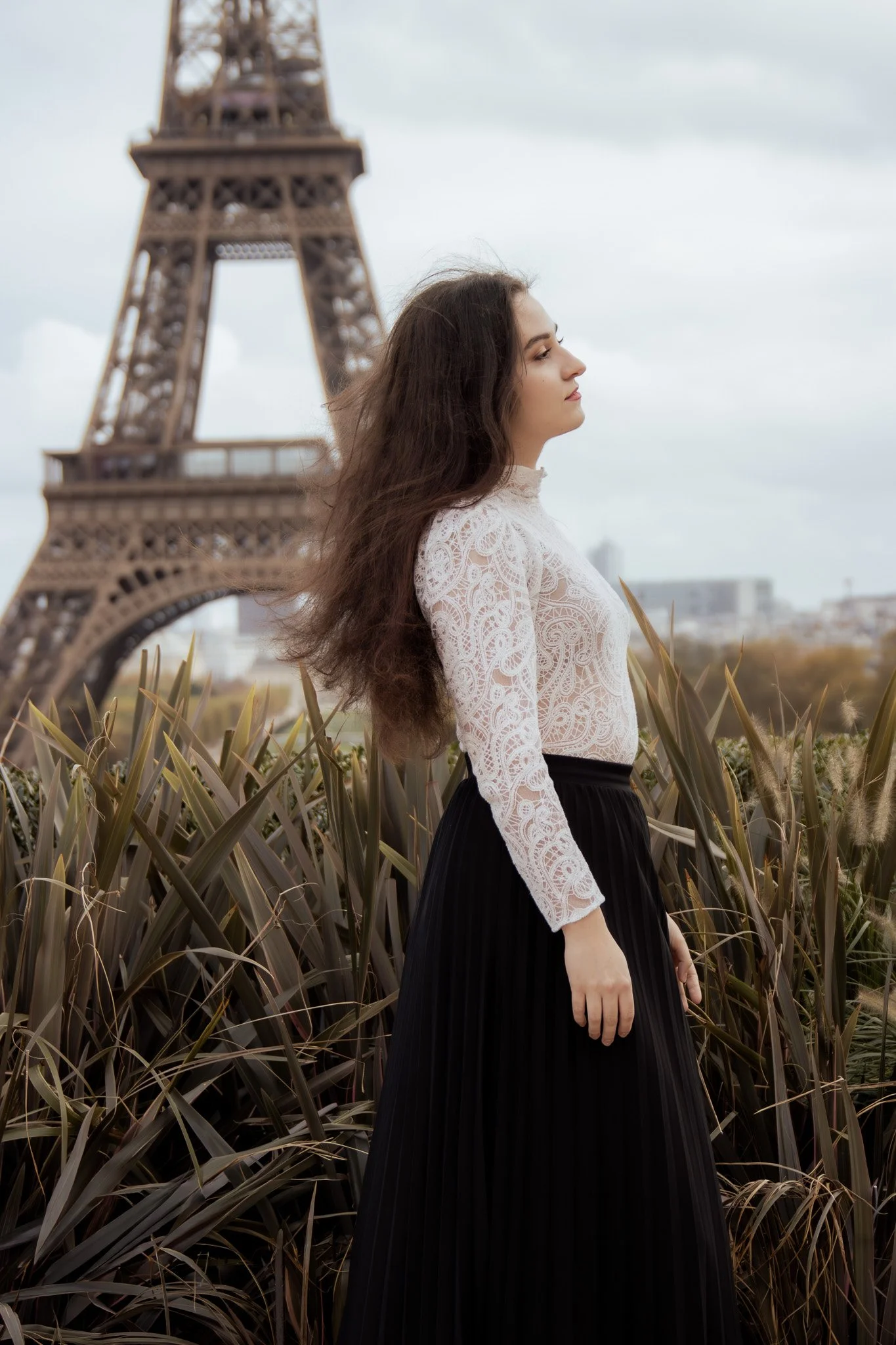 A woman in a white lace top and black pleated skirt standing in front of tall grass with the Eiffel Tower in the background.