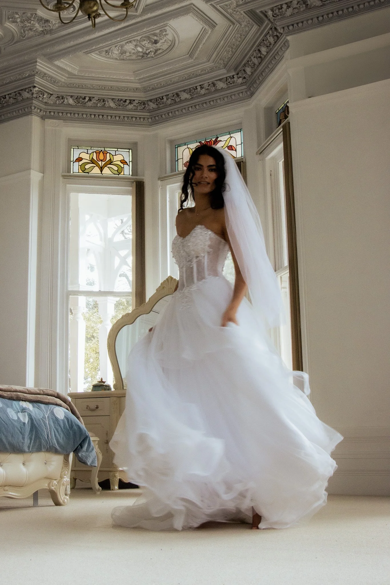 A bride in a white wedding gown and veil standing in a room with ornate ceiling details, large windows, and stained glass accents.