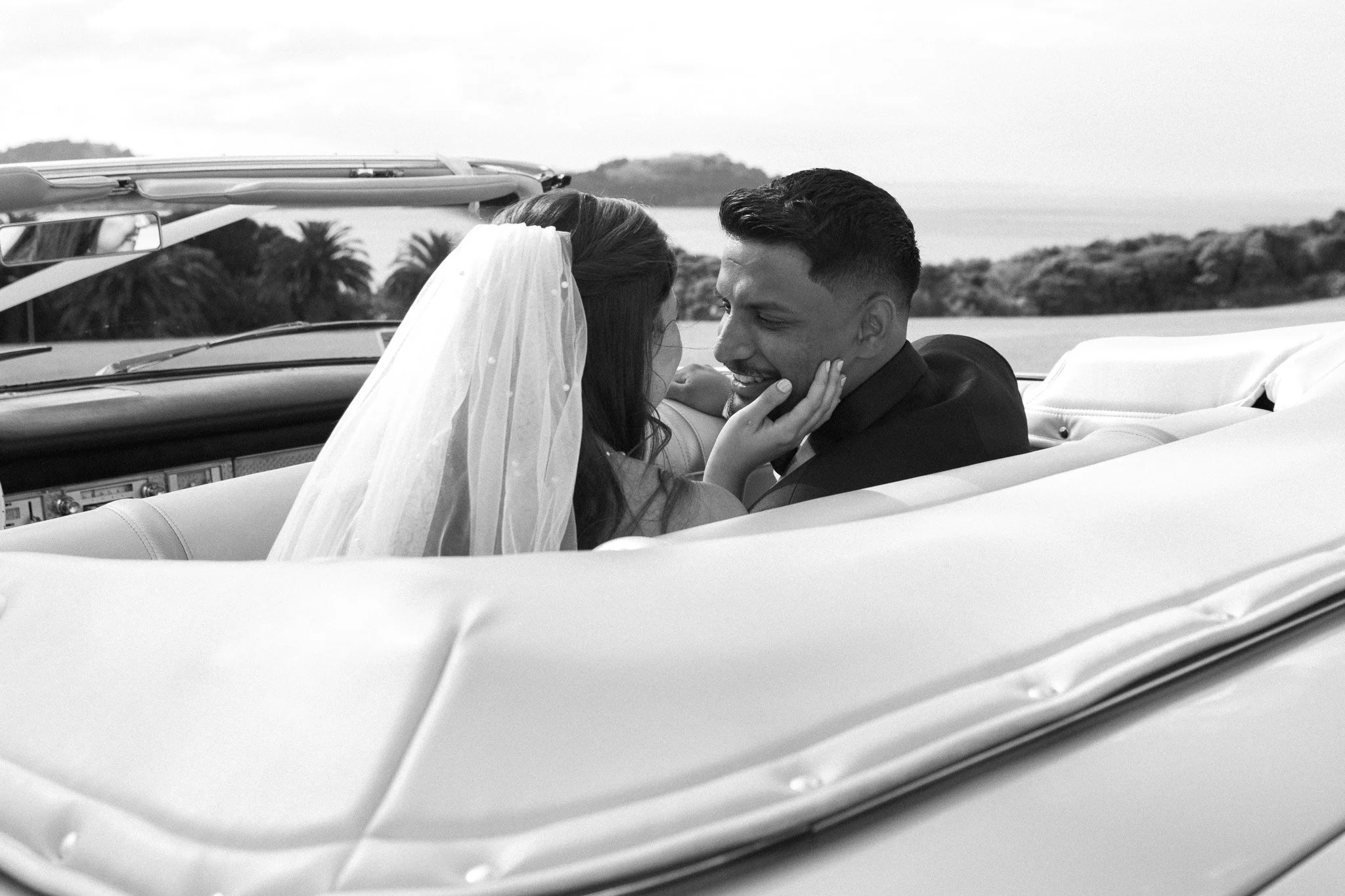A bride and groom sitting in a classic convertible car, facing each other affectionately, with a scenic background of hills and palm trees.