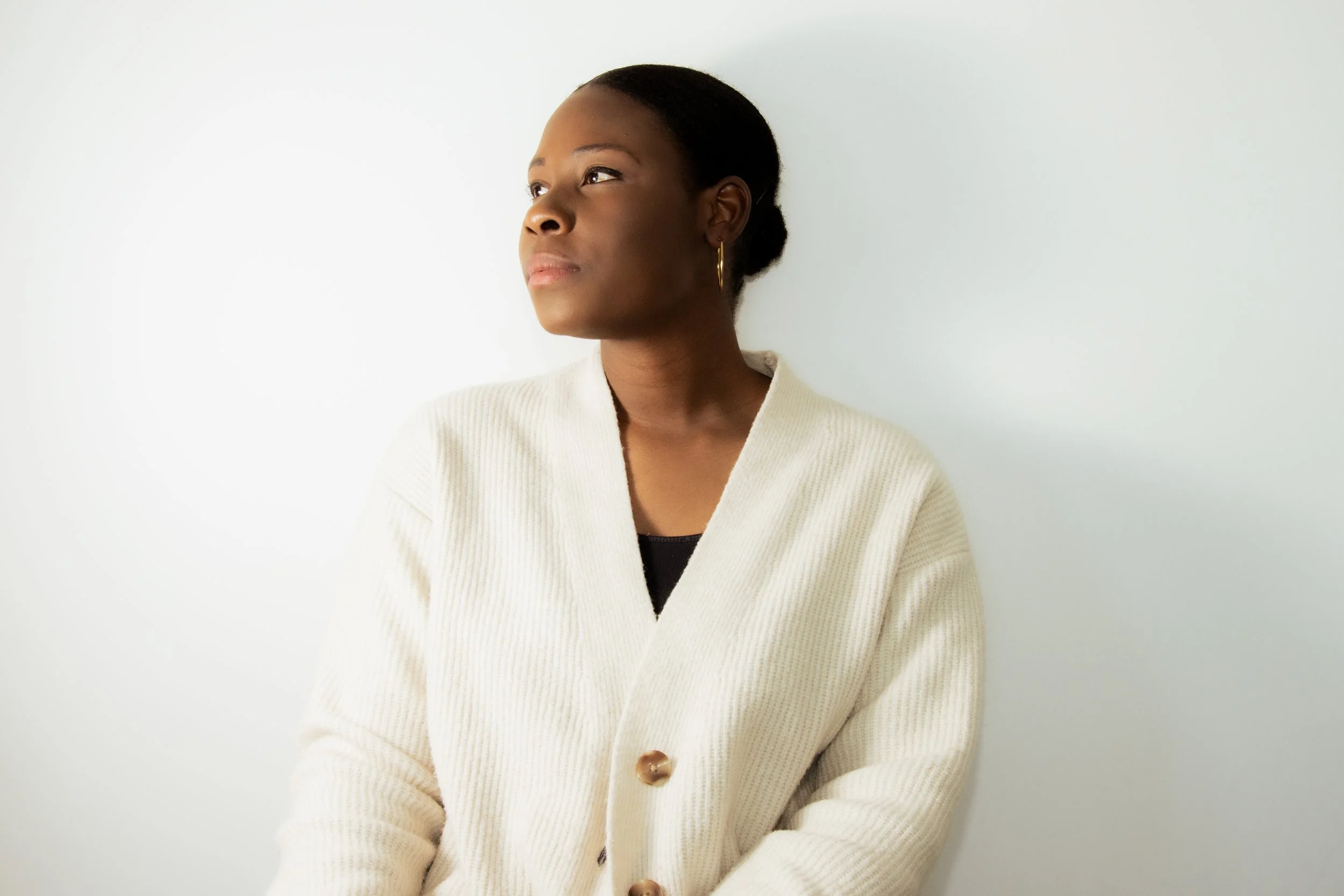 A woman with dark skin and black hair styled in a bun, wearing a cream-colored cardigan and gold hoop earrings, looking to the left against a white wall.