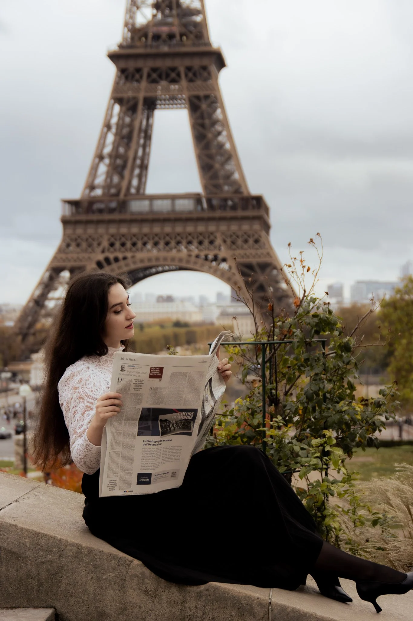 A woman with long dark hair, wearing a white lace top and black skirt, is sitting on a stone ledge reading a newspaper. The Eiffel Tower is in the background under an overcast sky, in Paris, France.