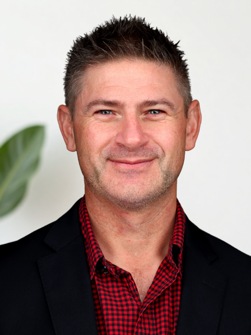 A man with short brown hair and light stubble, smiling, wearing a black blazer over a red and black checkered shirt, against a white background with a green plant visible on the left.