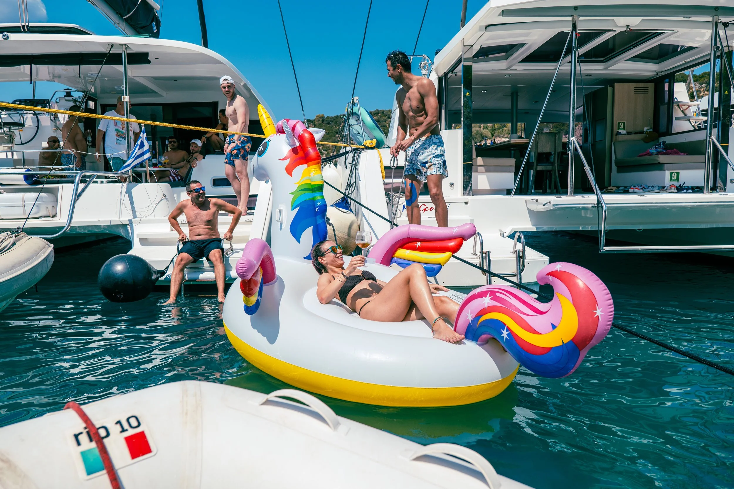 Aerial view of a small island with green vegetation surrounded by turquoise water, with several anchored sailboats nearby, and larger islands and mountains in the distance under clear blue sky.