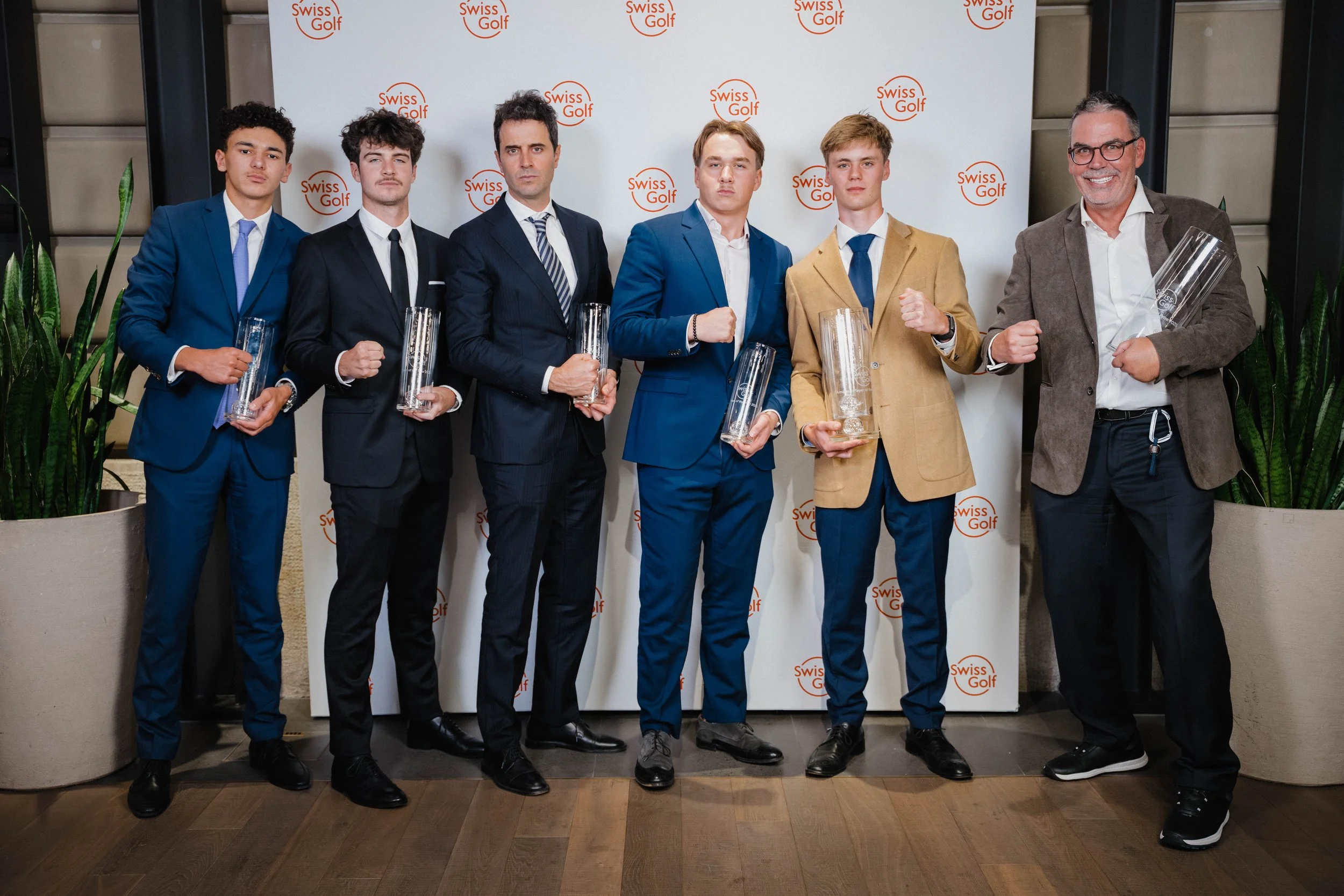 Seven young men in suits holding trophies, posing for a photo at a Swiss Golf event, with a backdrop displaying the Swiss Golf logo, flanked by two large plants.