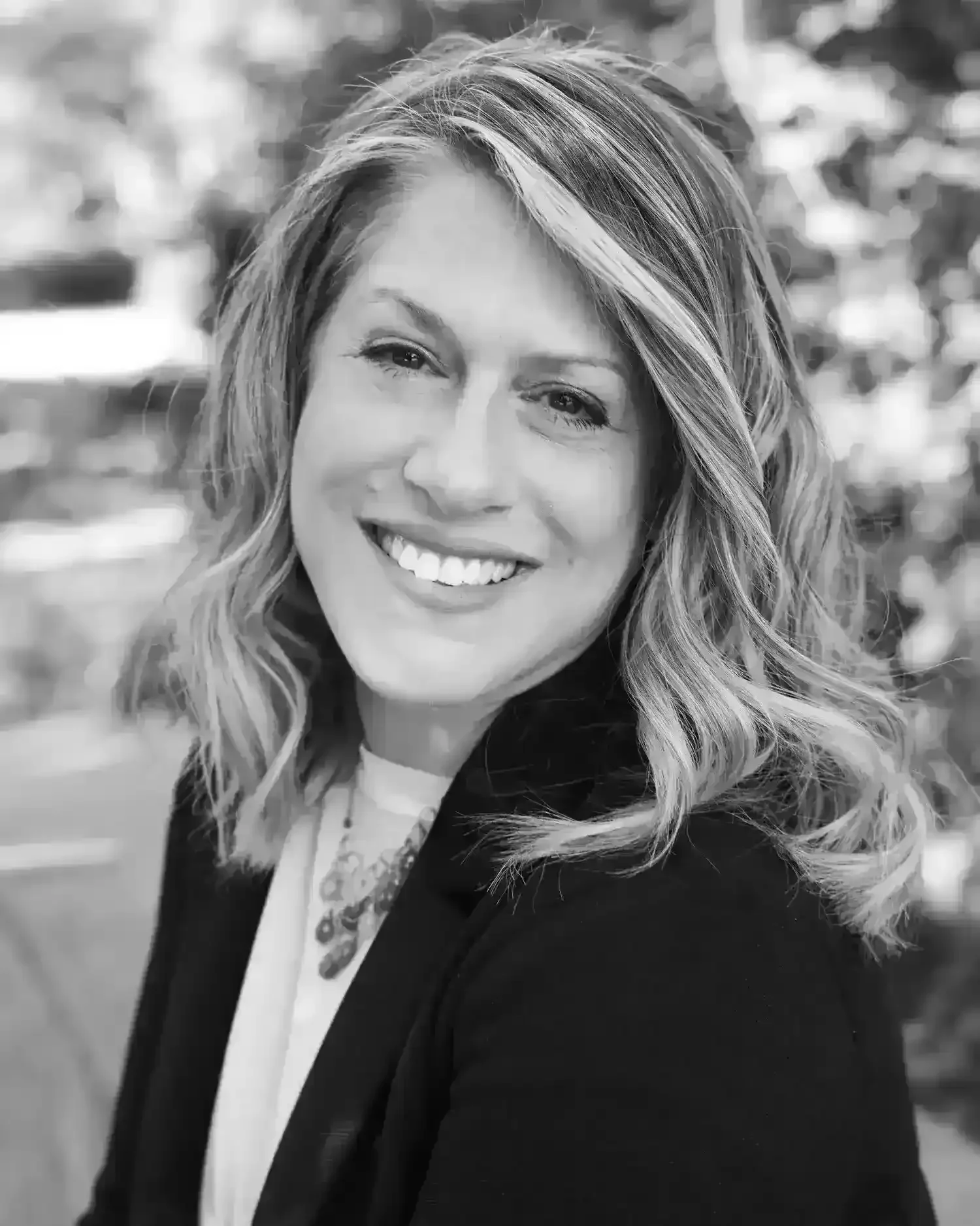Black and white portrait of a smiling woman with wavy shoulder-length hair, wearing a blazer and a decorative necklace, outdoors with blurred background.