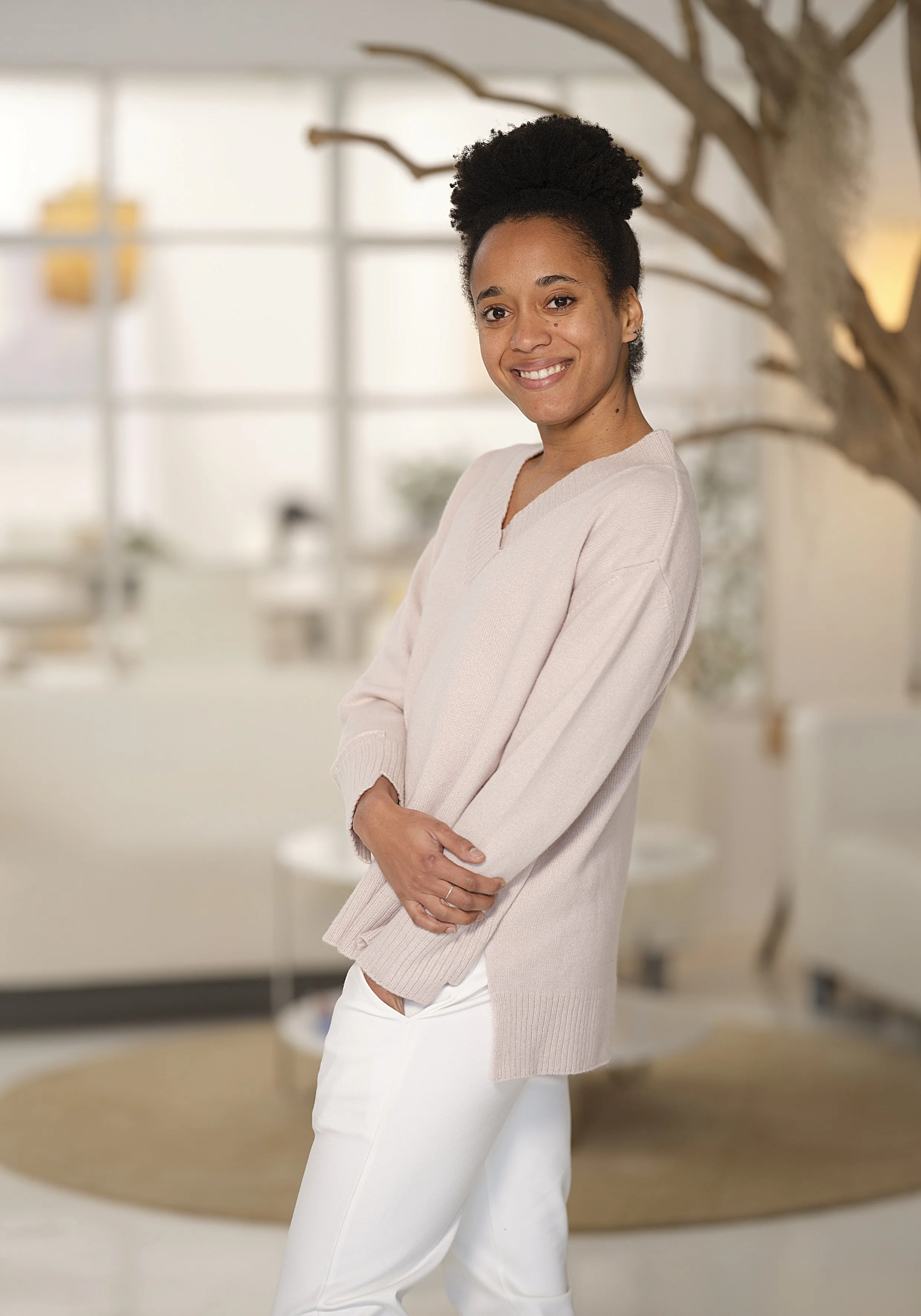 A smiling woman with curly hair styled in an updo, wearing a light pink sweater and white pants, stands indoors with her arms crossed. The background features large windows, a tree branch, and a blurred interior setting.